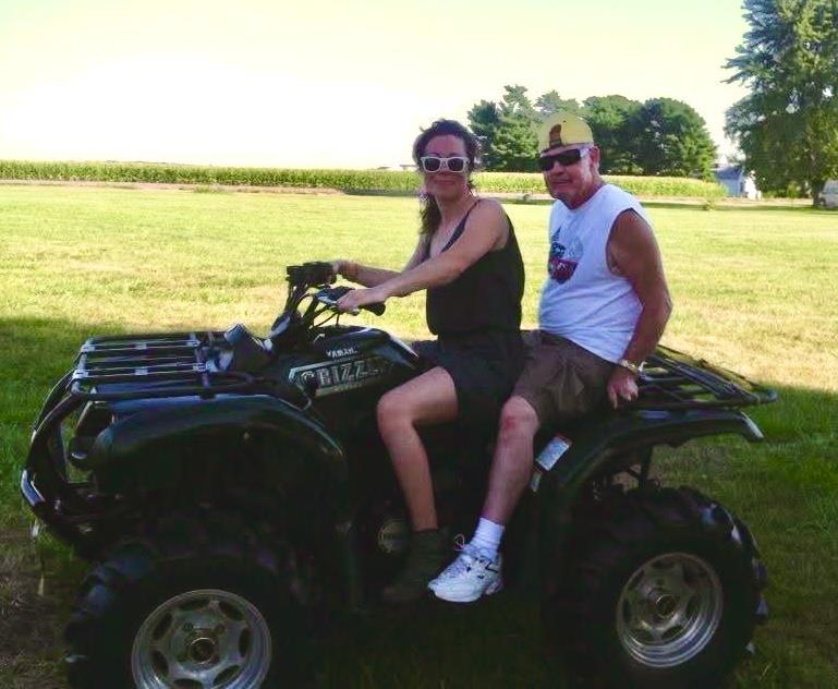 A man and woman confidently ride an ATV together across a vibrant green field under clear skies.