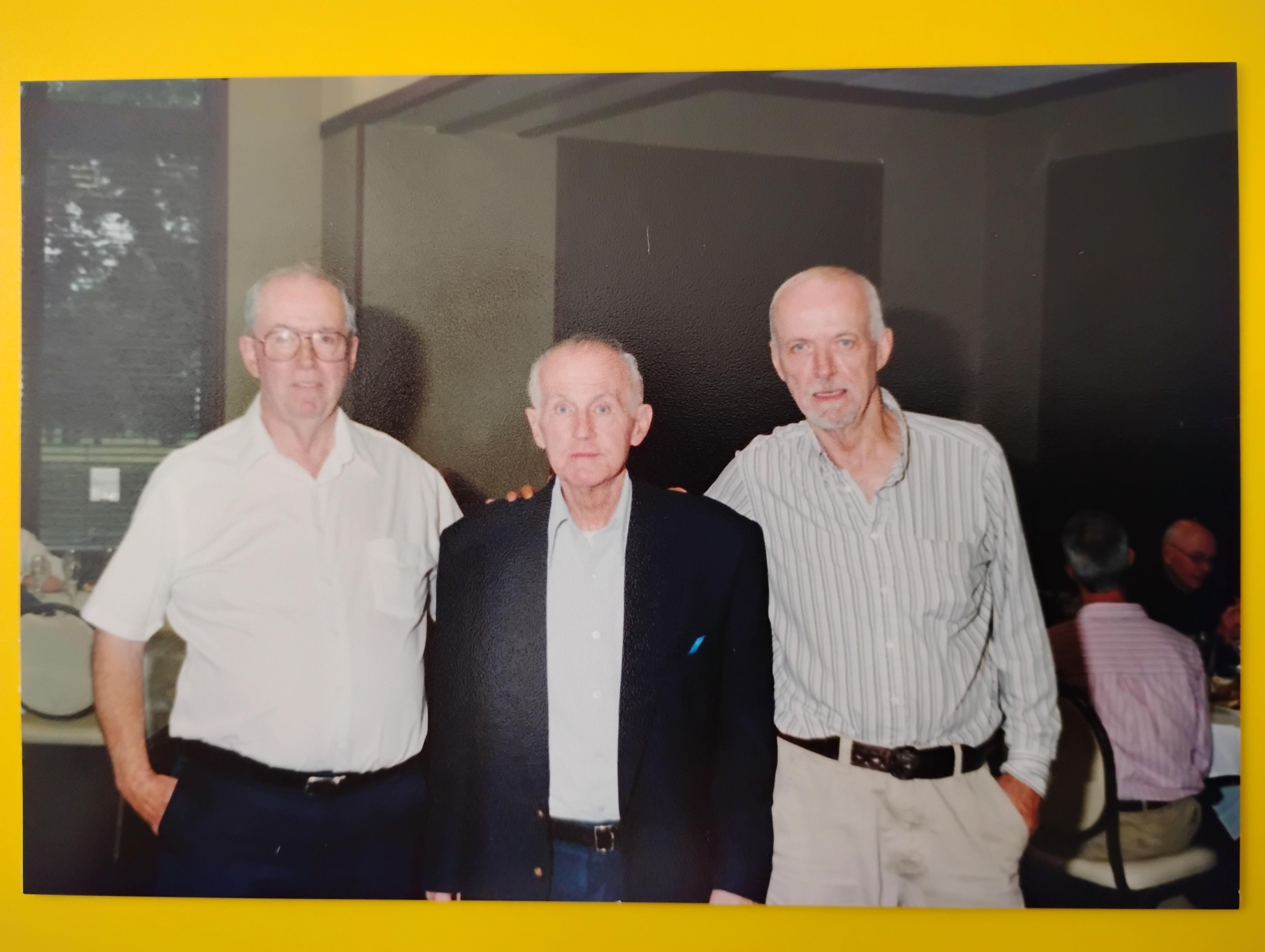 Three elderly men gather together, smiling warmly, in a casual indoor setting during a reunion.