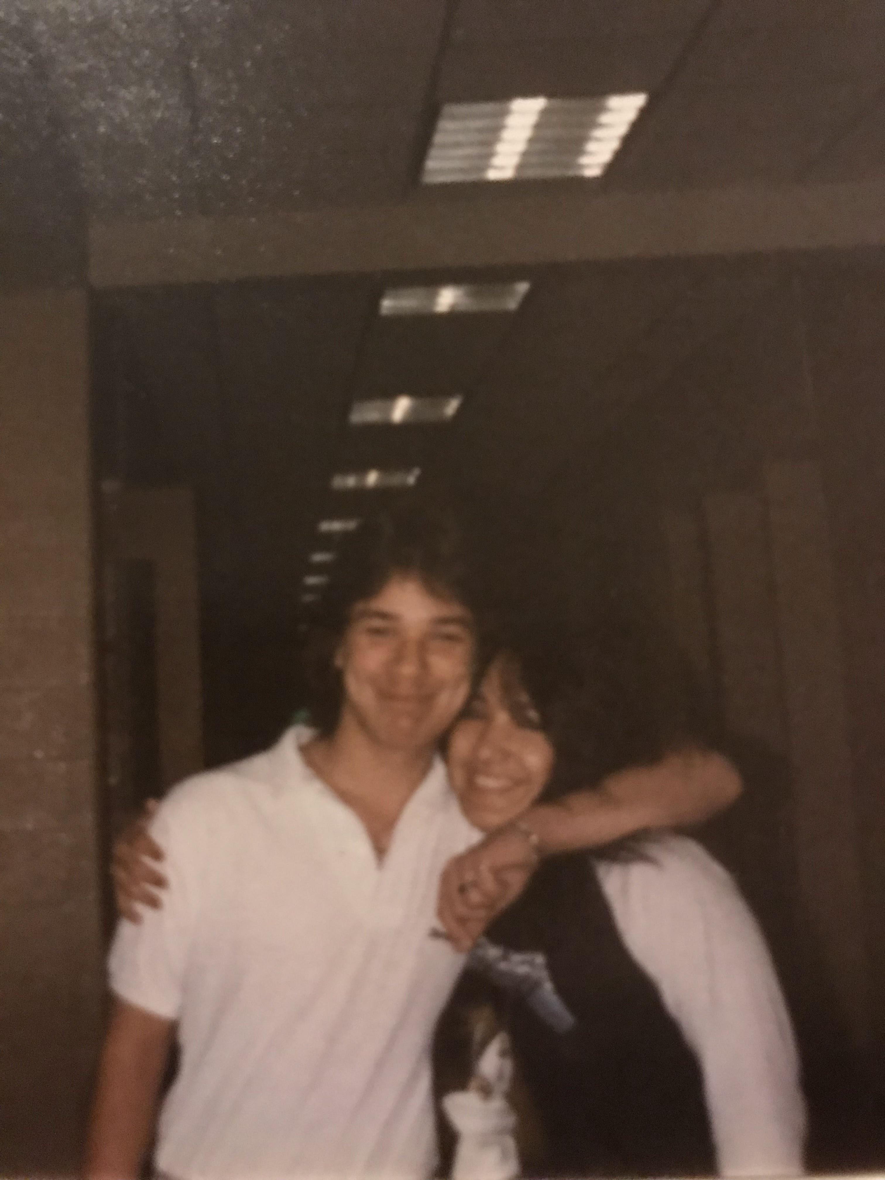 Two friends smile and embrace in a hallway filled with warm lighting during a fun event.