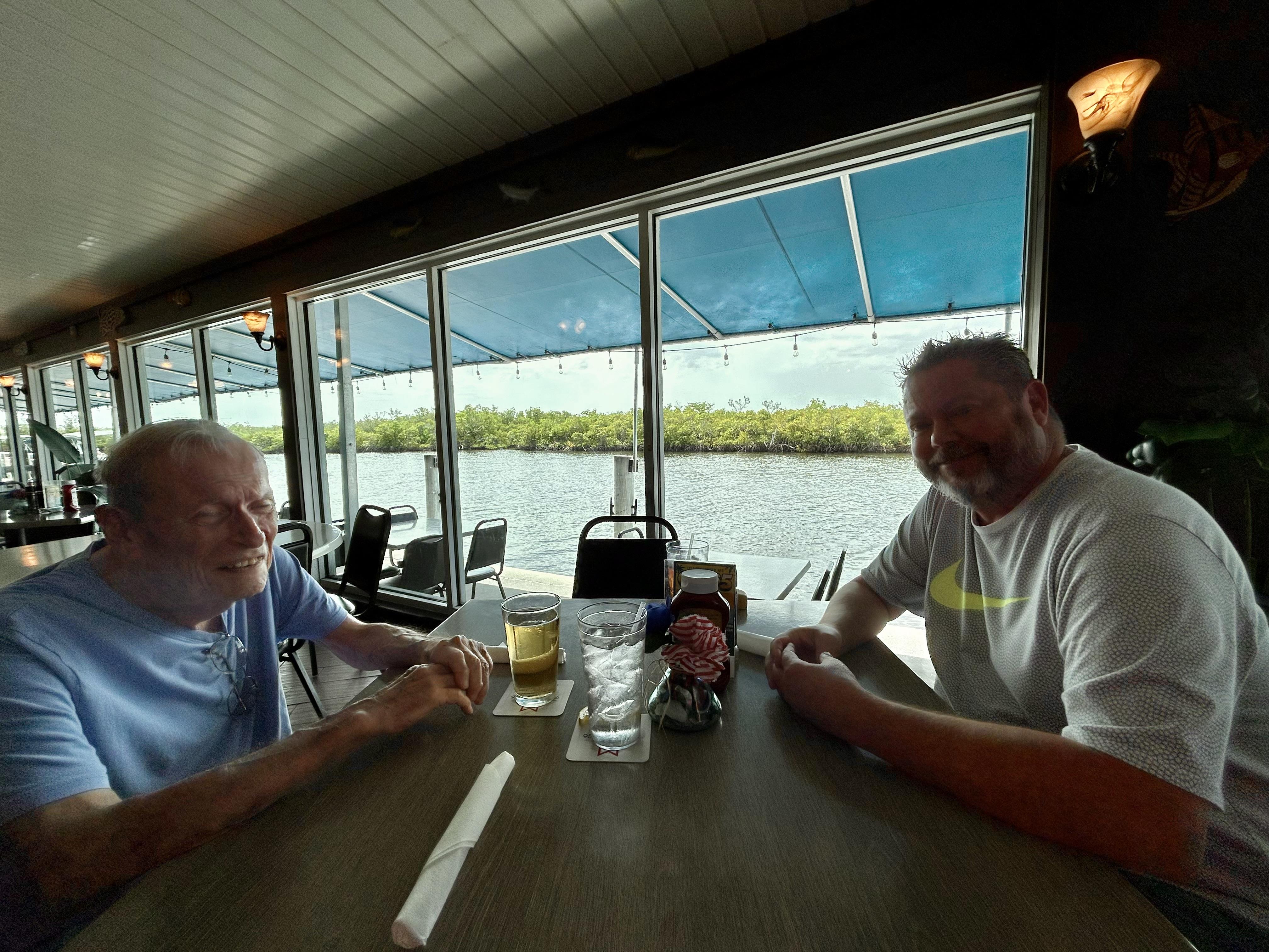 Two friends sit at a table by the water, enjoying drinks and conversation in bright sunlight.