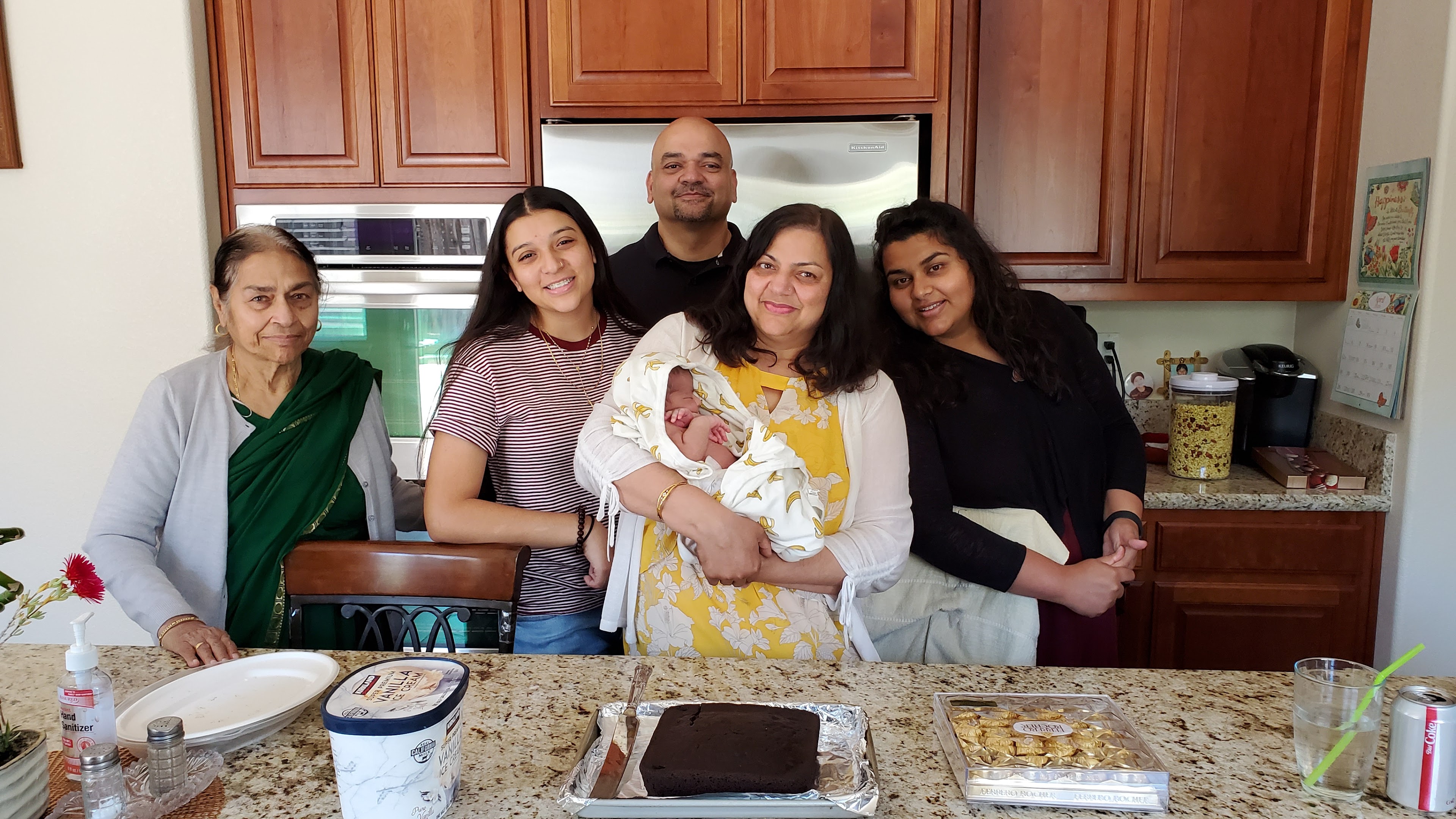 Family poses together in a kitchen, celebrating the arrival of a newborn with smiles and joy.