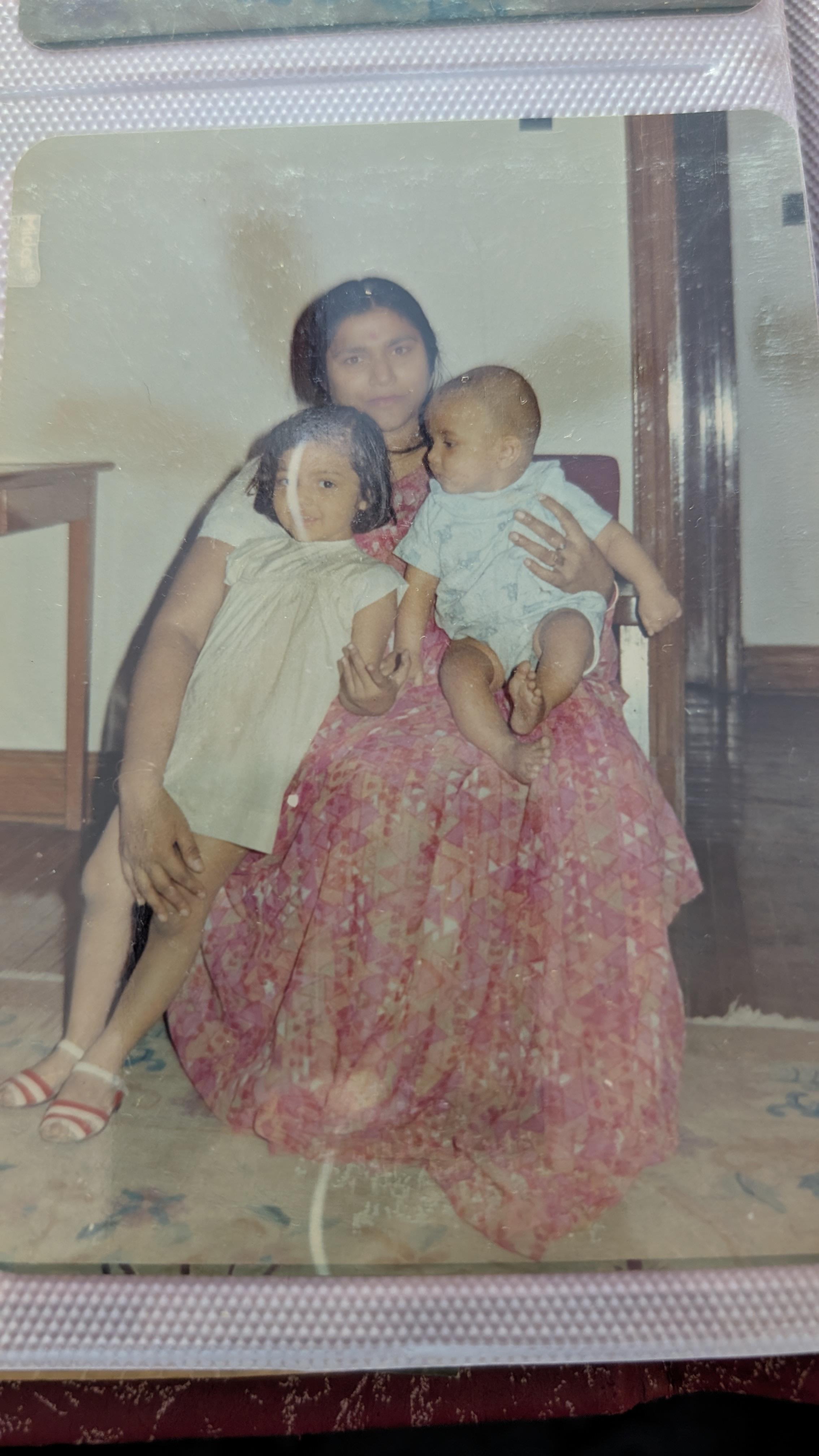 Woman in a floral dress holds a baby while a little girl sits nearby in a cozy indoor setting.