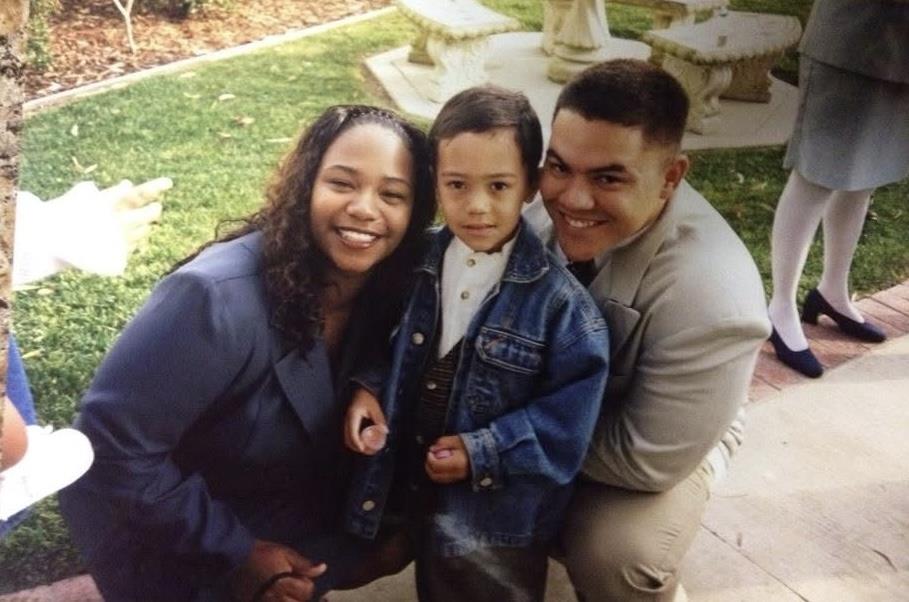 Two adults smile broadly while standing beside a young child in casual clothing outdoors.