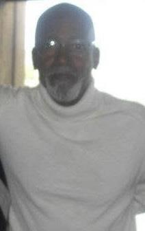 An elderly man sits comfortably indoors, wearing a white turtleneck and glasses, smiling warmly.