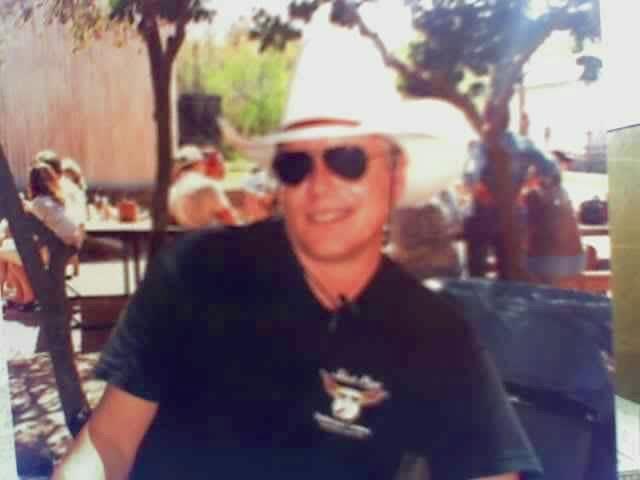 A man enjoys a sunny day at an outdoor cafe, smiling and wearing a stylish hat and sunglasses.