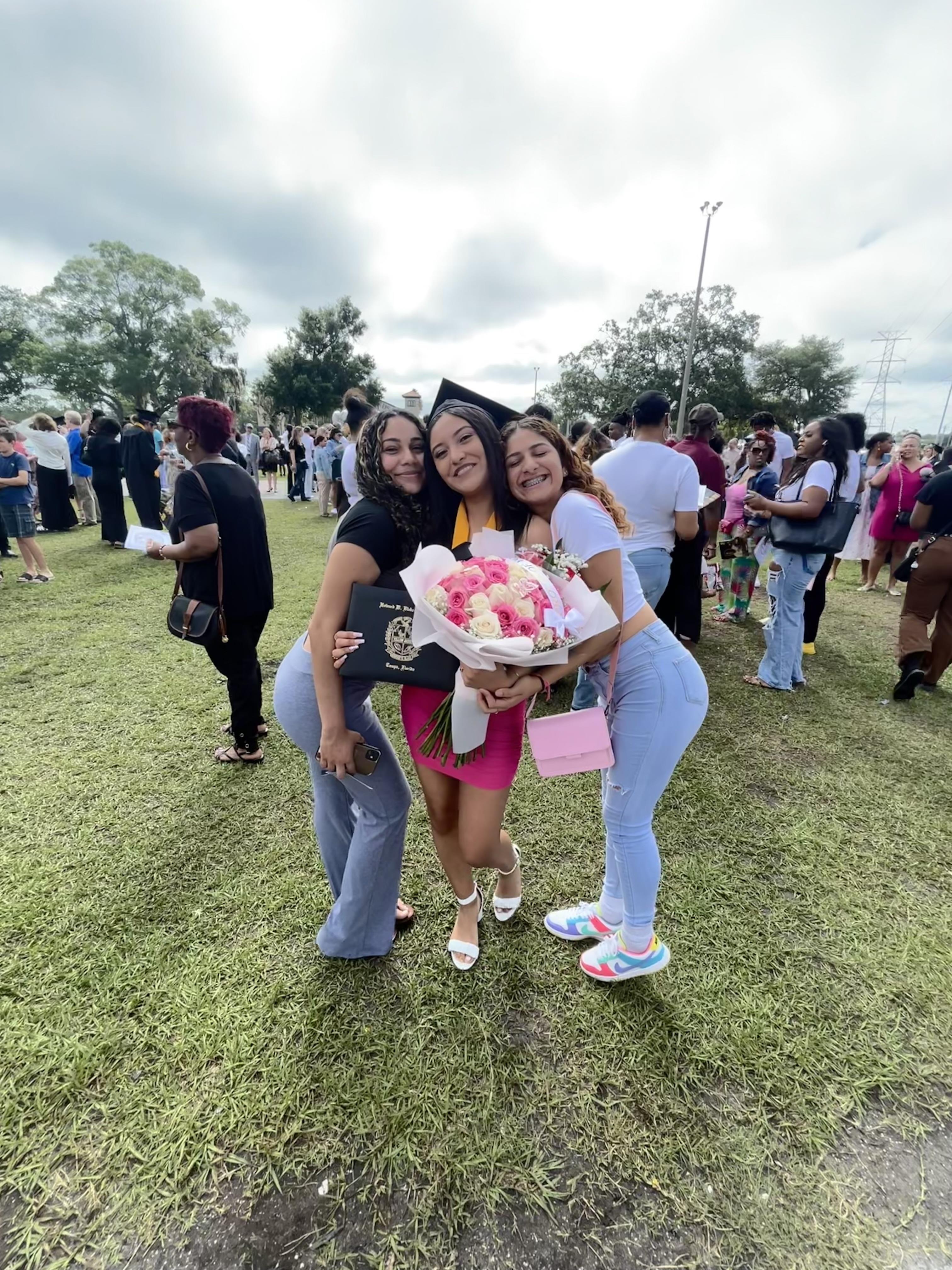 Three friends pose happily on graduation day while holding flowers in a grassy area.