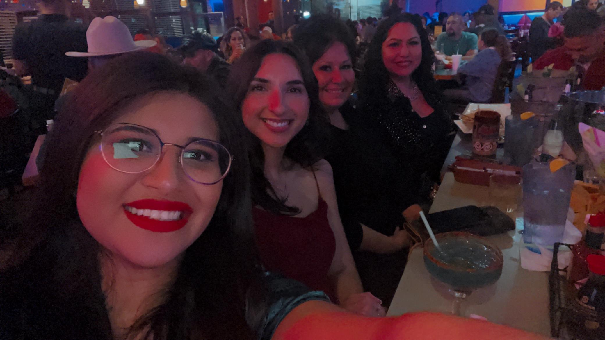 Four friends take a selfie, smiling and celebrating at a bustling restaurant at night.