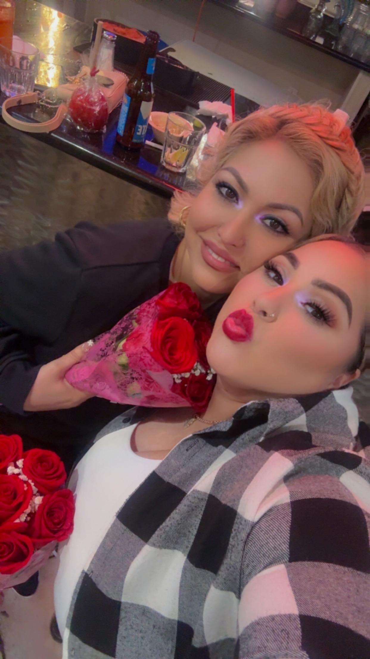 Two friends pose together, holding bouquets of red roses in a lively cafe atmosphere.