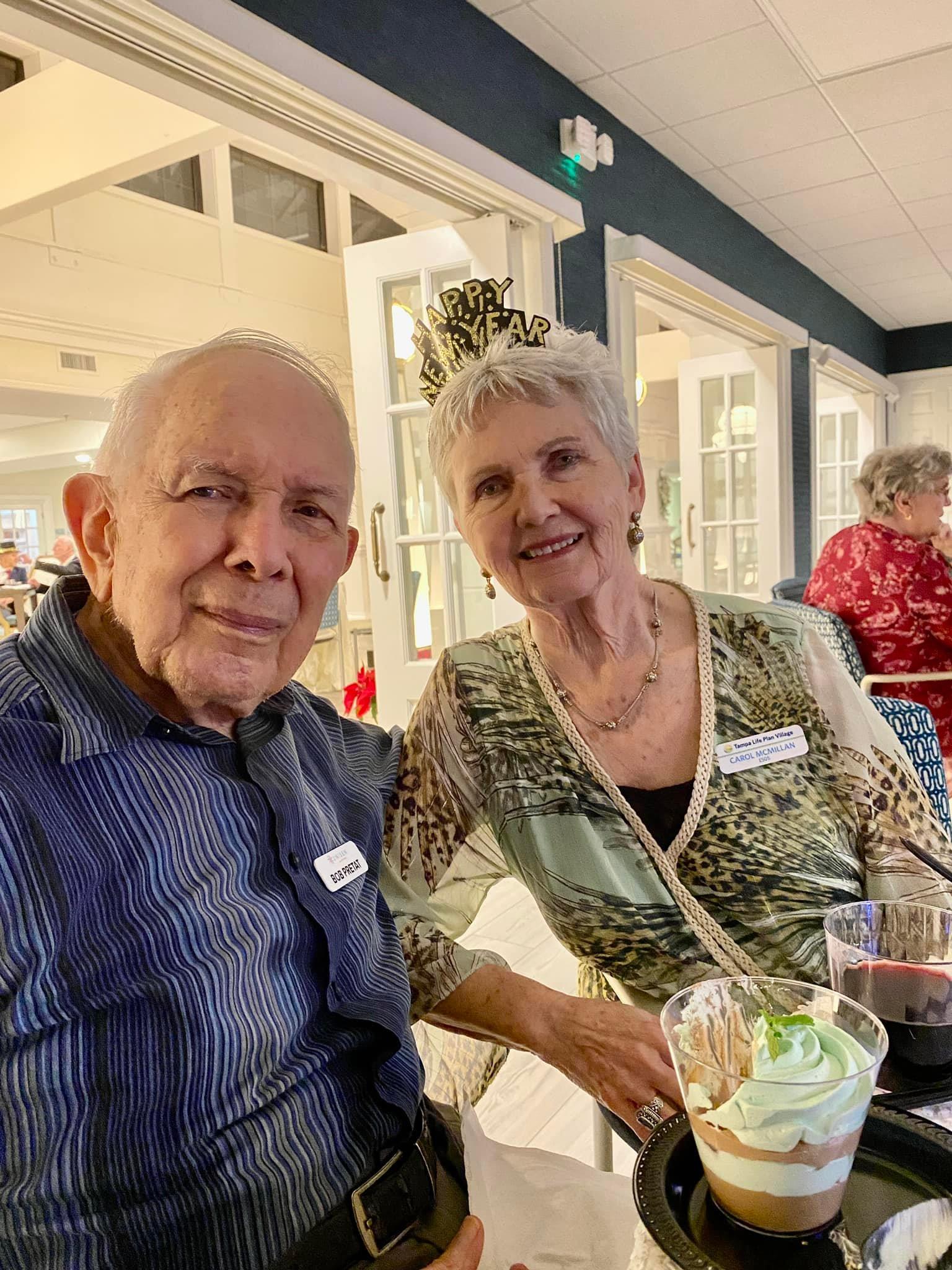 An elderly couple enjoys drinks and smiles together at a lively retirement home event.