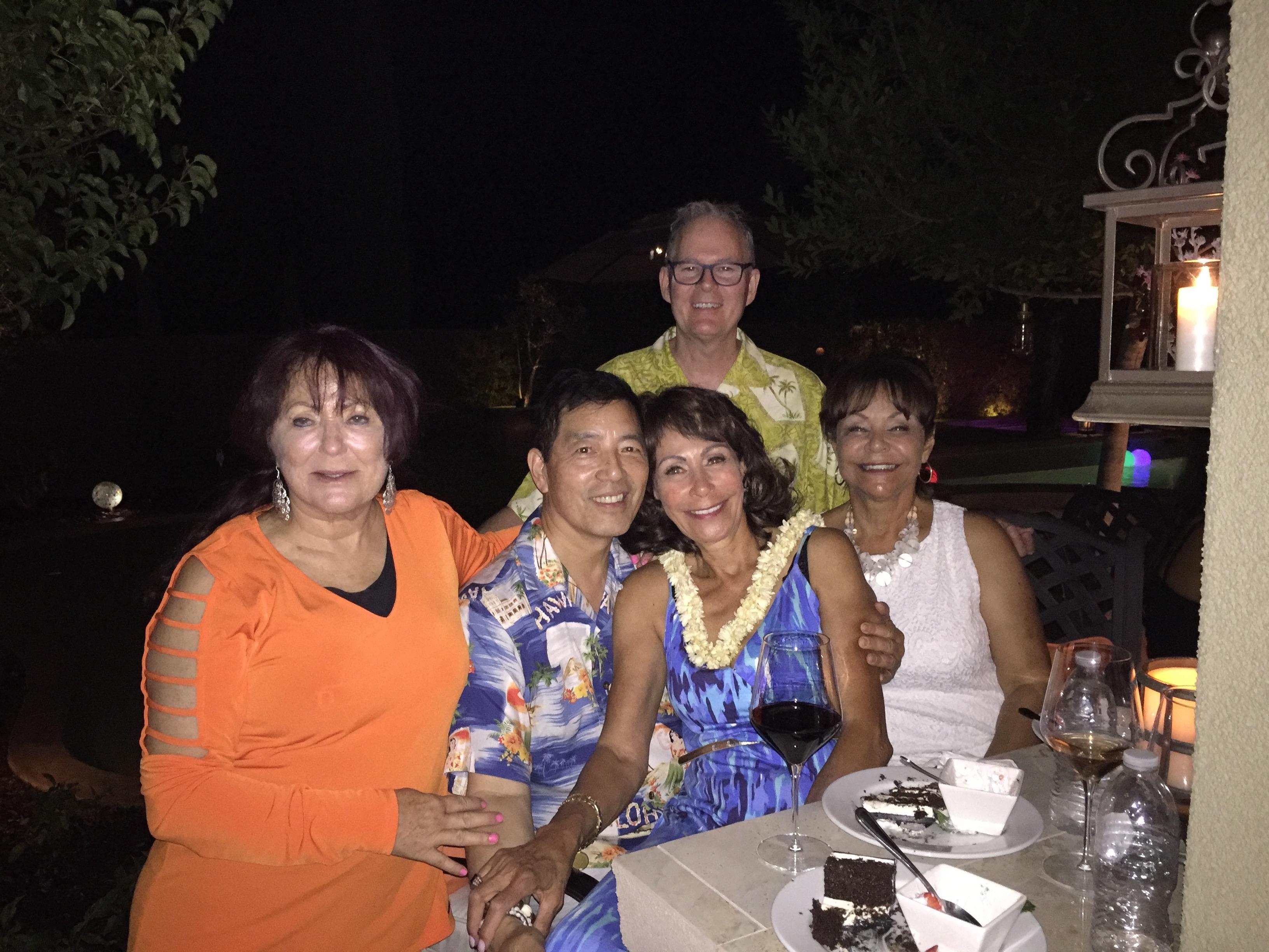 Friends gather around a table under the night sky, celebrating with laughter and drinks.