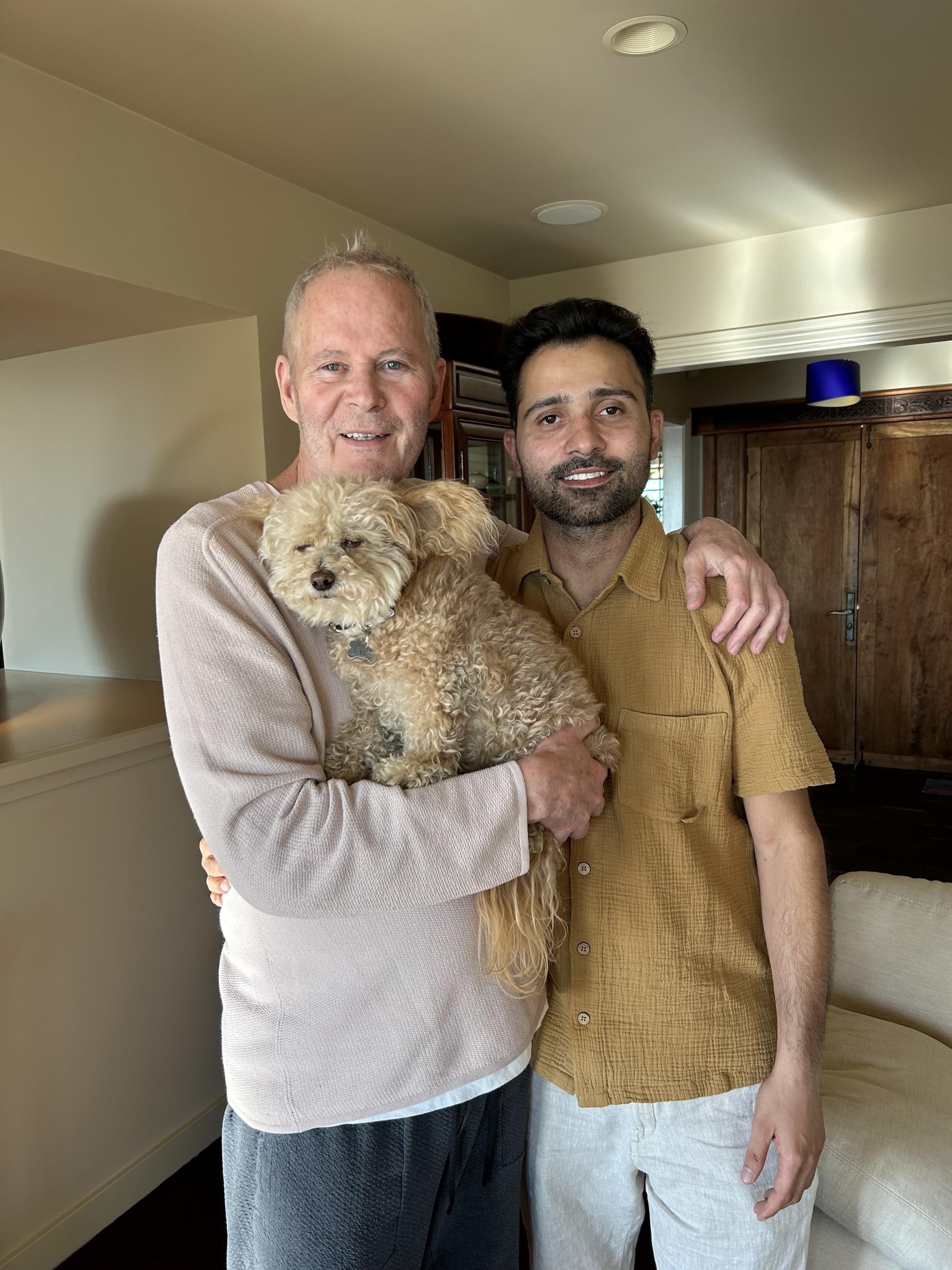 Two friends pose happily in a welcoming living room, holding a small dog and smiling.