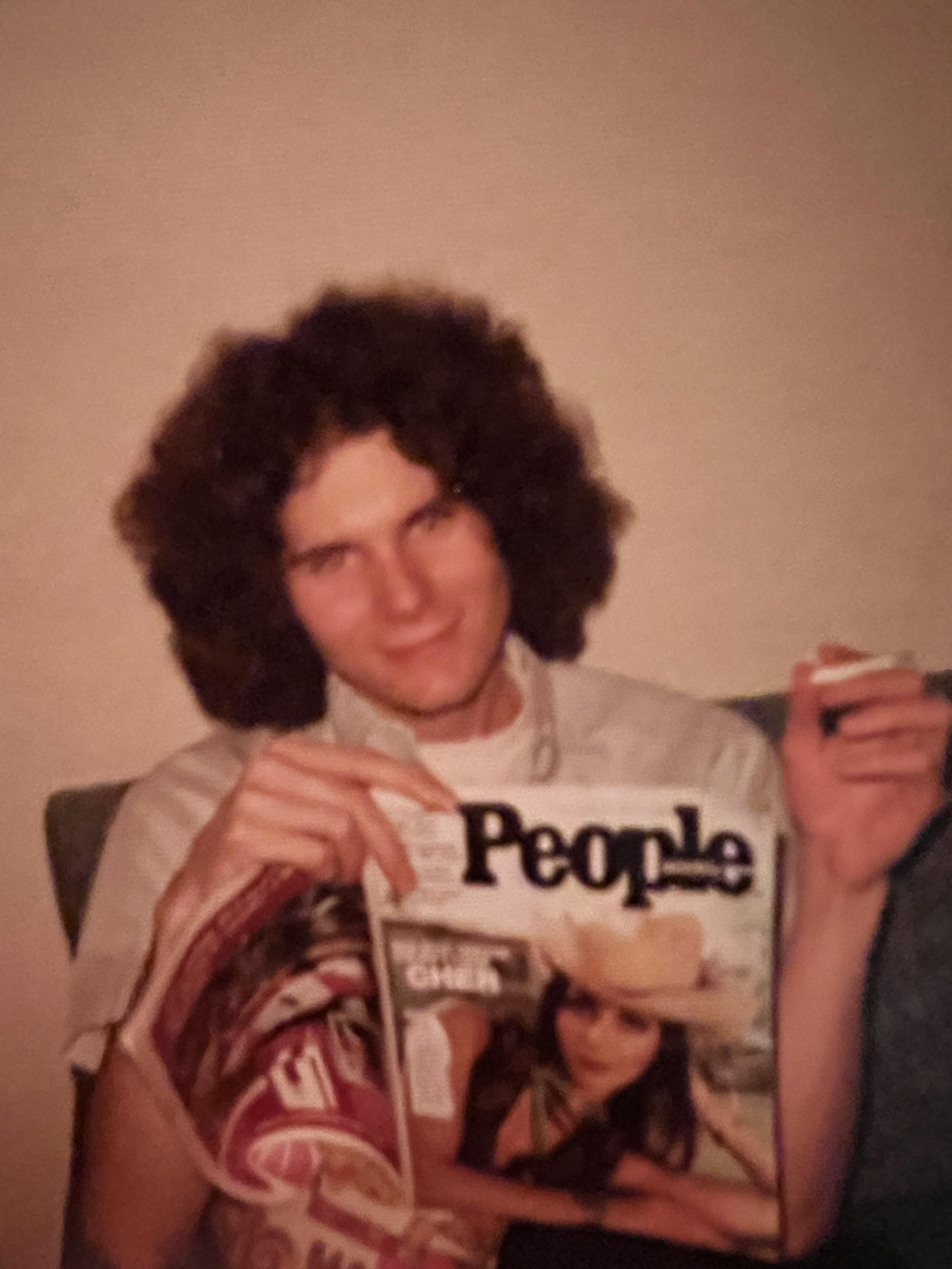 A young man with curly hair sits on a couch holding a magazine while smoking a cigarette.