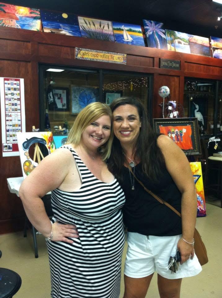 Two friends smile and pose for a picture at an art gallery filled with colorful artworks.