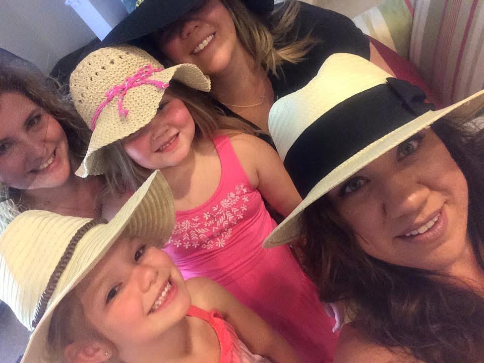 Women and young girls smile together, wearing colorful summer hats at a lively indoor setting.