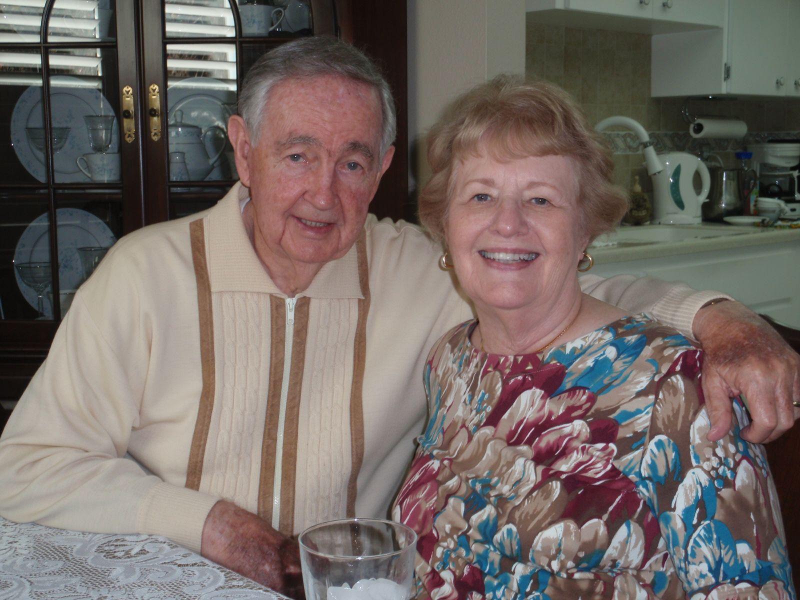 An elderly couple smiles happily at the camera, sharing a loving moment in their home.