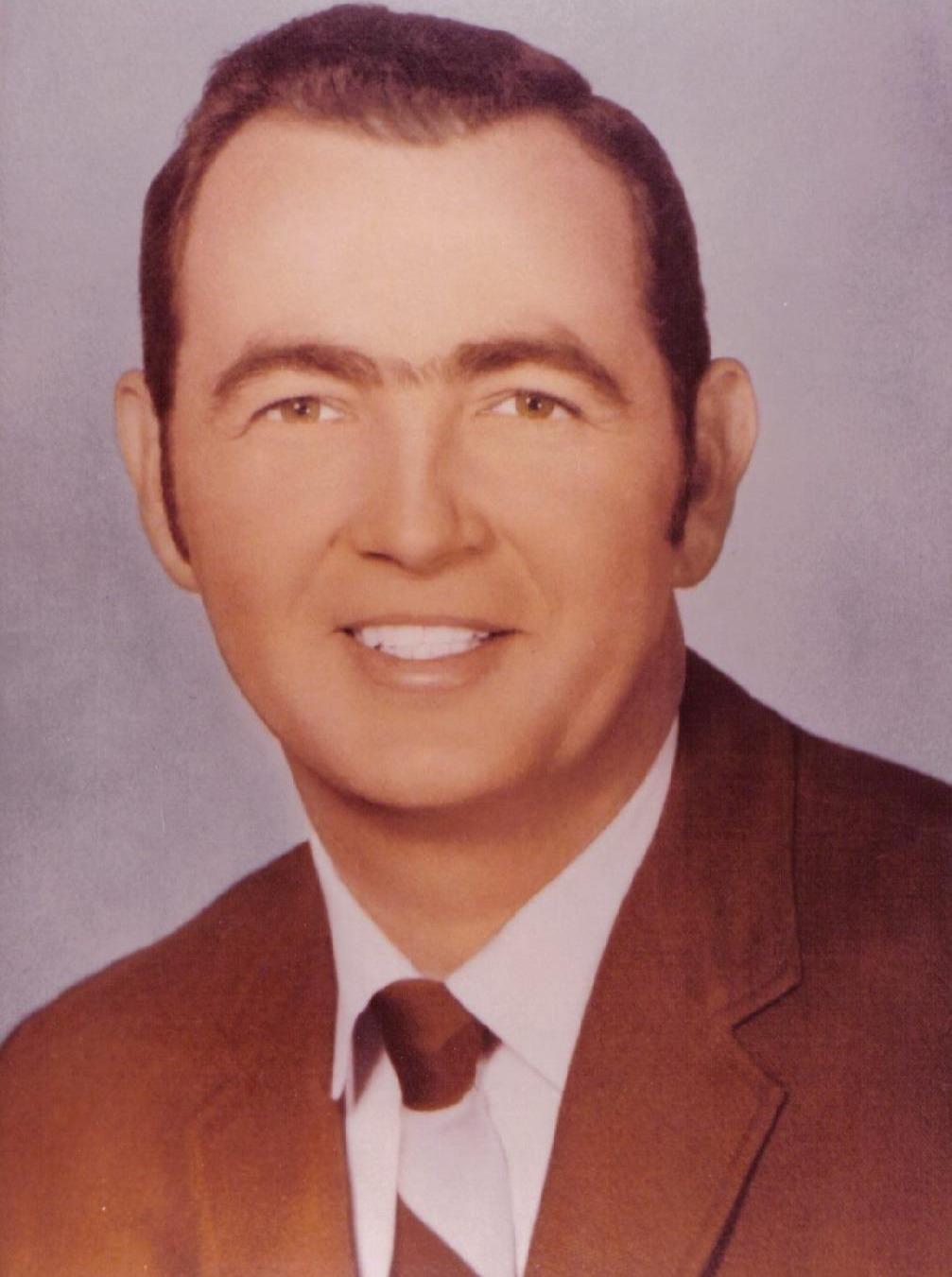 A man wearing a brown suit and tie smiles warmly for a formal portrait indoors.