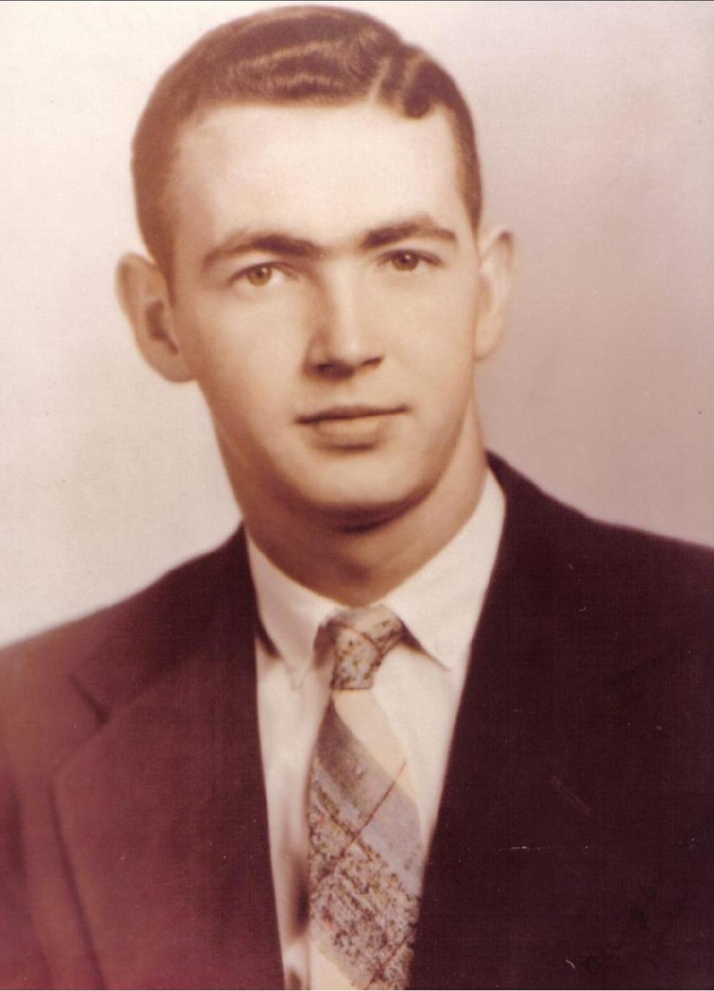 A young man with short hair smiles confidently in a suit and patterned tie, circa 1950s.
