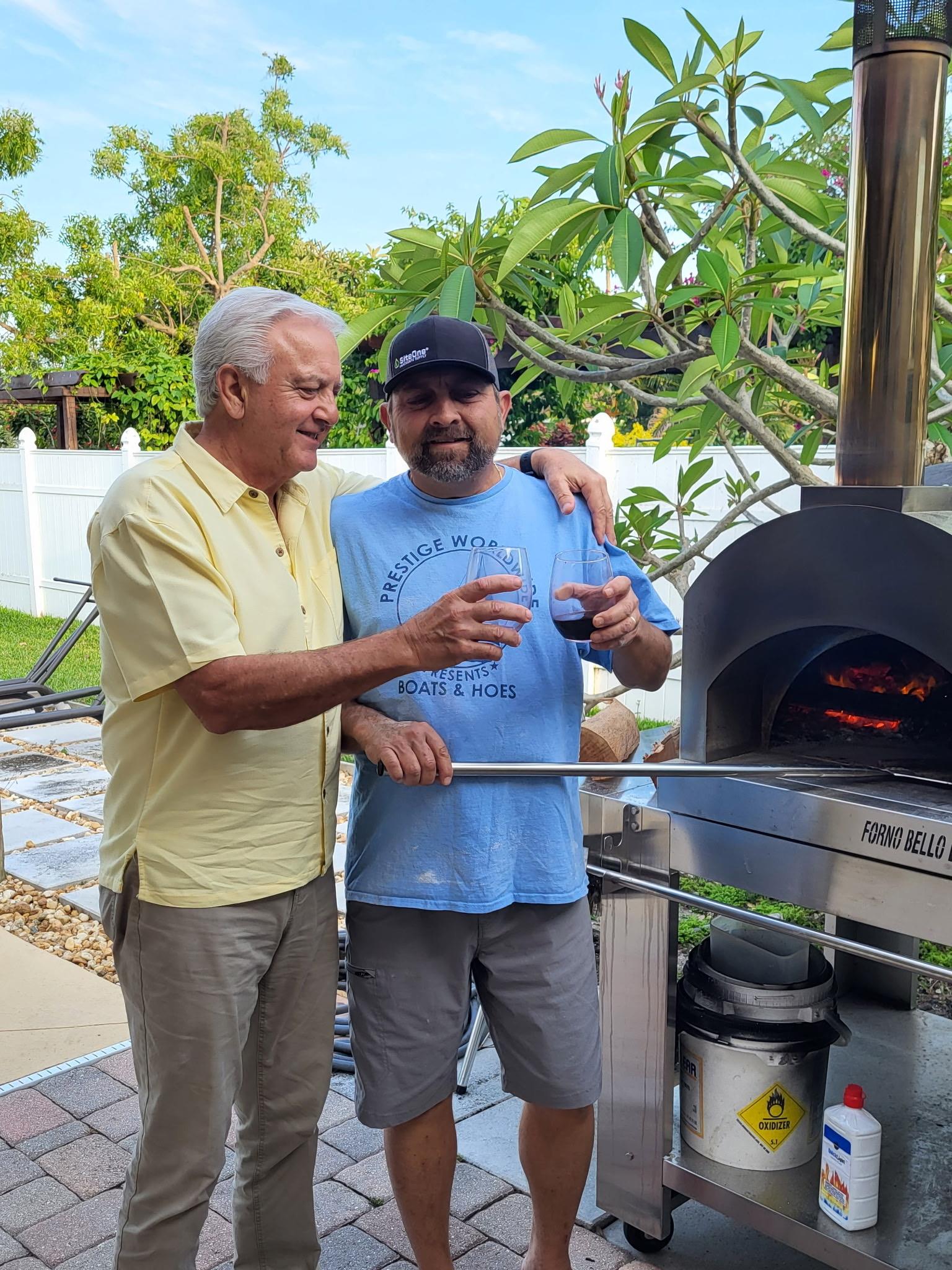 Two friends raise their drinks and smile while cooking in a backyard pizza oven.