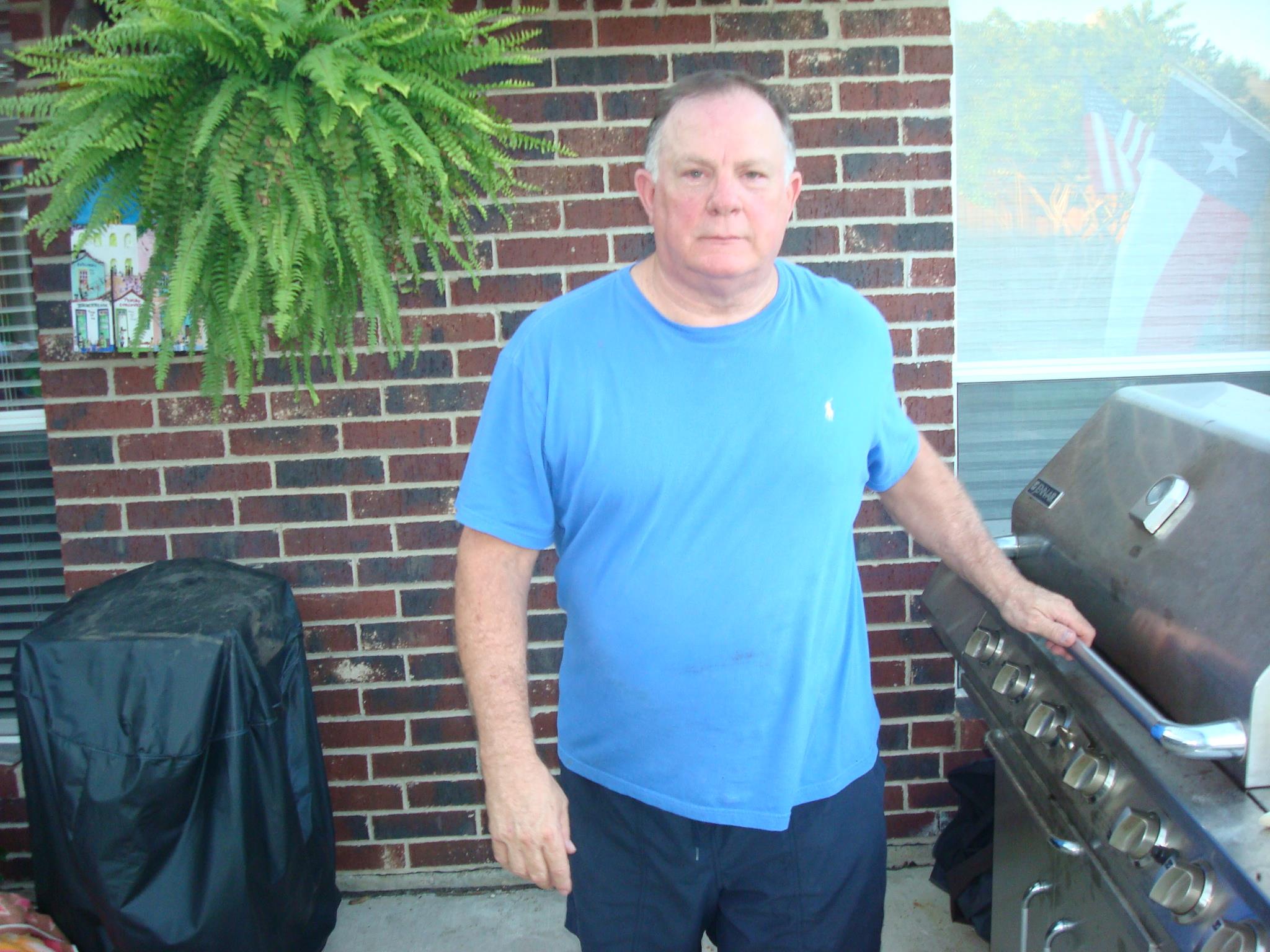 Man stands by a grill with a relaxed expression, surrounded by greenery and brick wall.