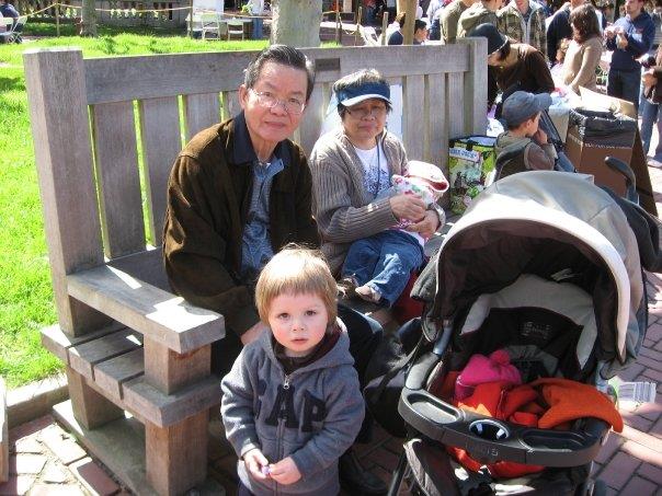 A man and a woman rest on a bench while a child stands in front during a busy day in the park.