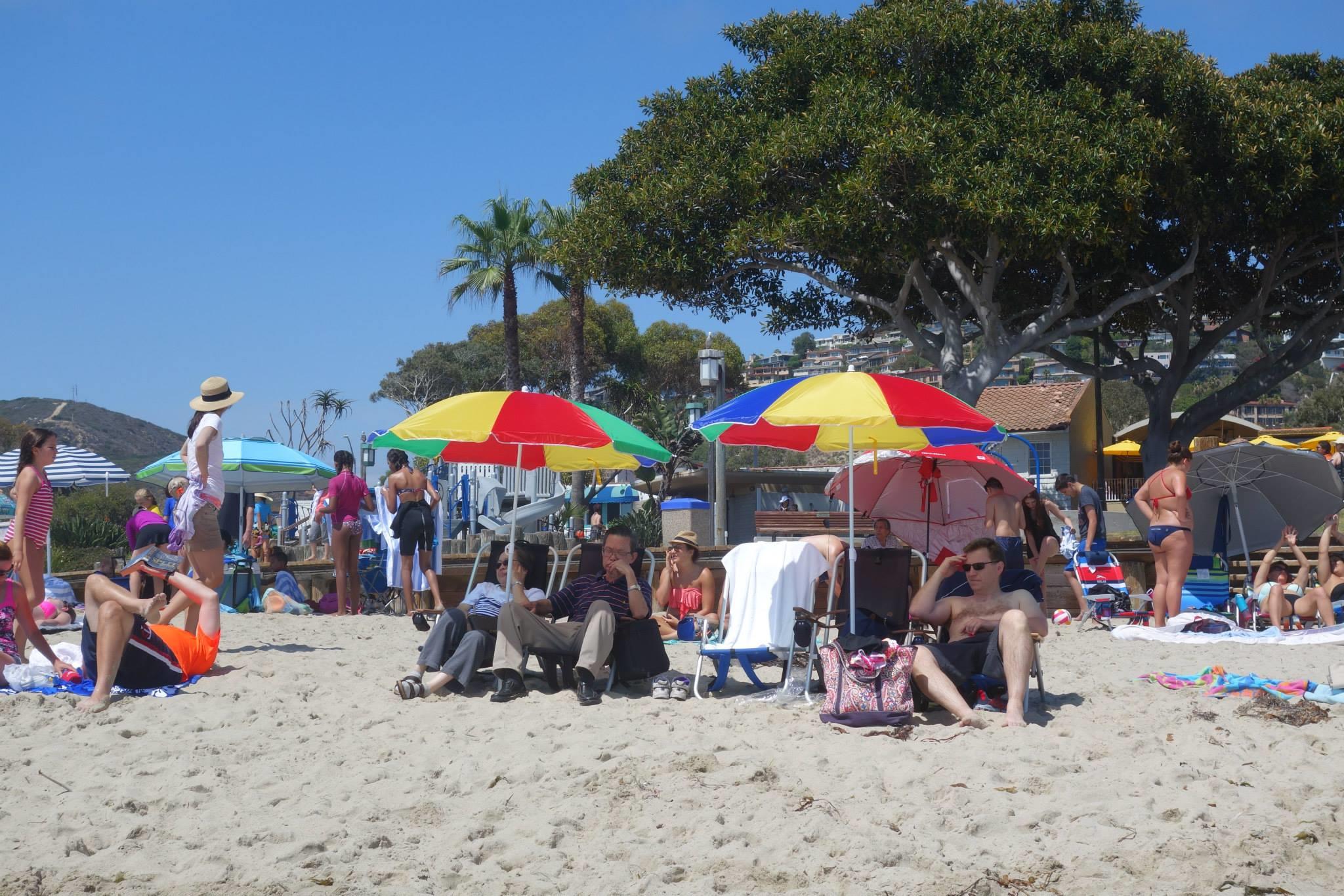 People sit on the sand at the beach enjoying the sun under large umbrellas as others walk by.