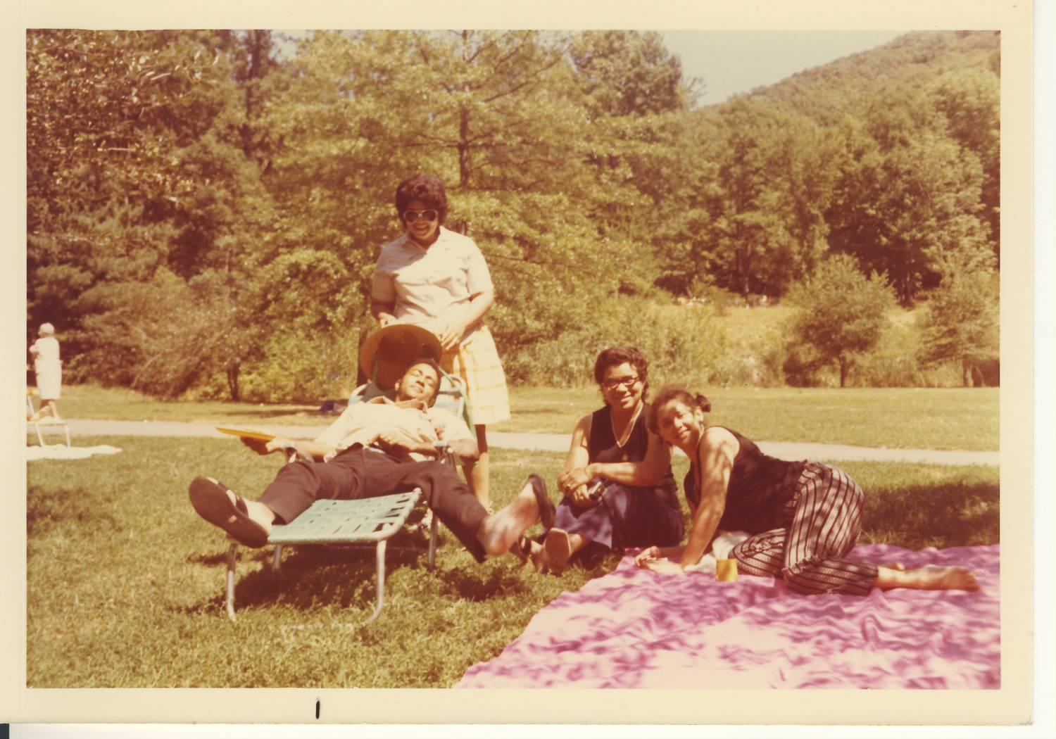 Five friends relax on a colorful blanket under the sun in a peaceful park surrounded by trees.