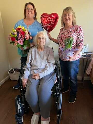 Two smiling women present flowers and a balloon to an elderly woman in a wheelchair.