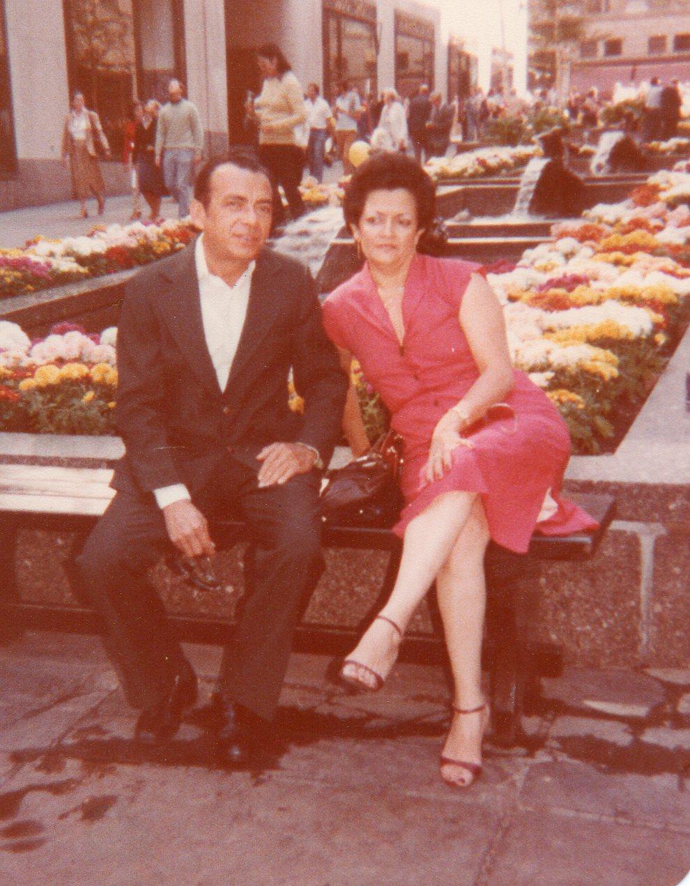 A couple relaxes on a bench surrounded by colorful flowers in a lively park.