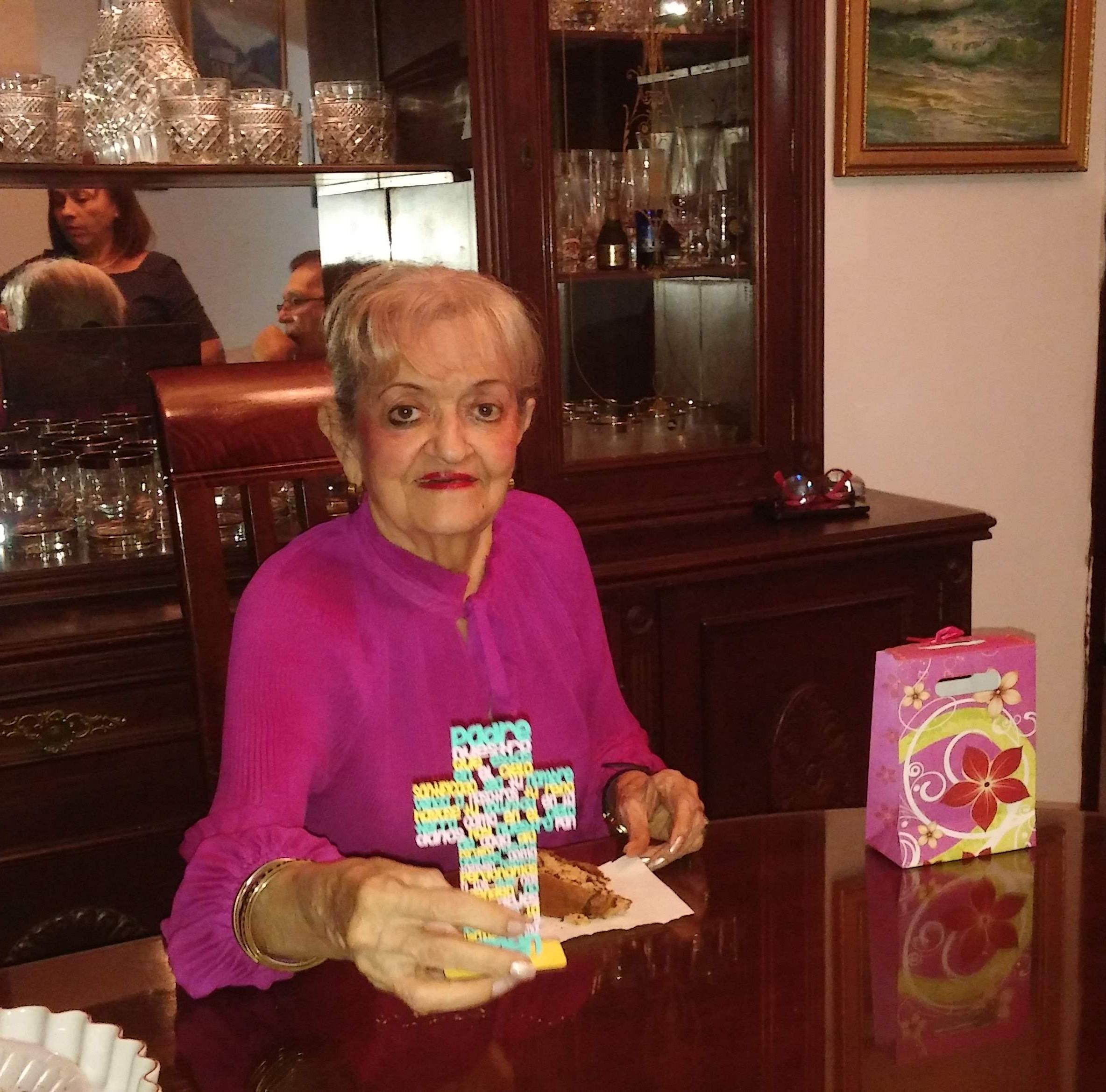 An elderly woman enjoys a moment at a polished wooden table, holding a colorful cross.