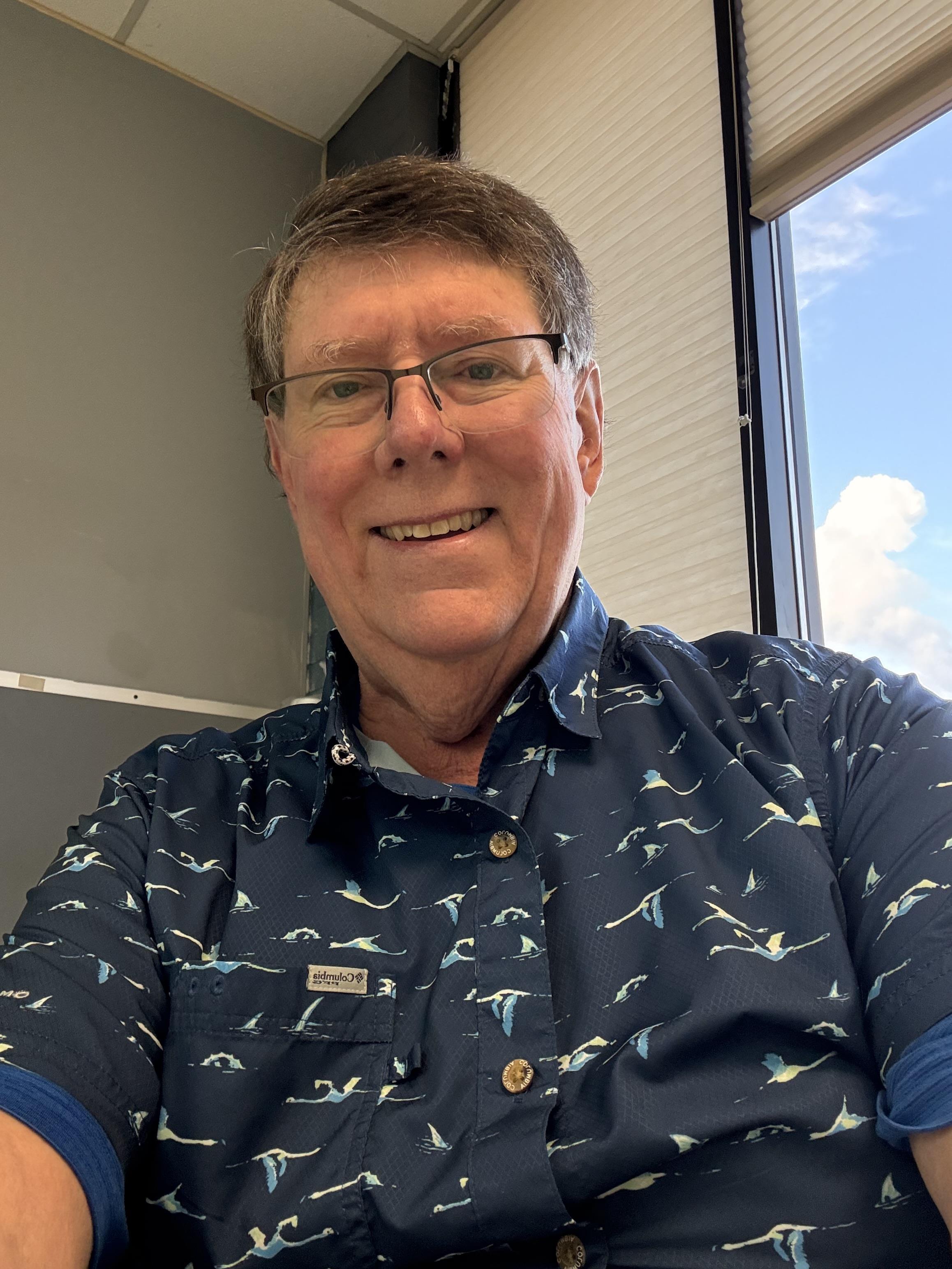 A man with glasses smiles while seated indoors, sunlight shining through a window.