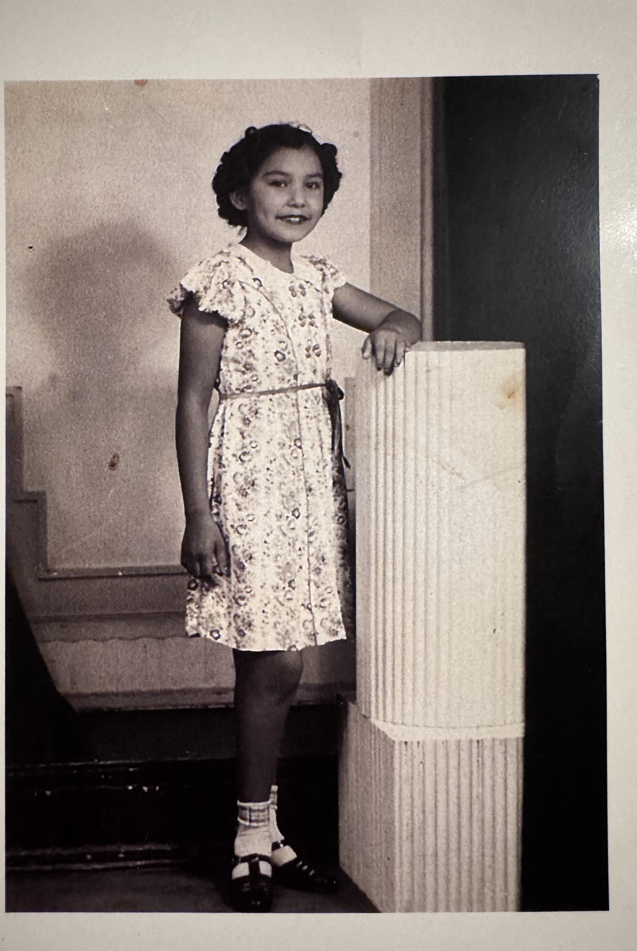 A girl stands beside a decorative column, wearing a styled dress and smiling warmly.