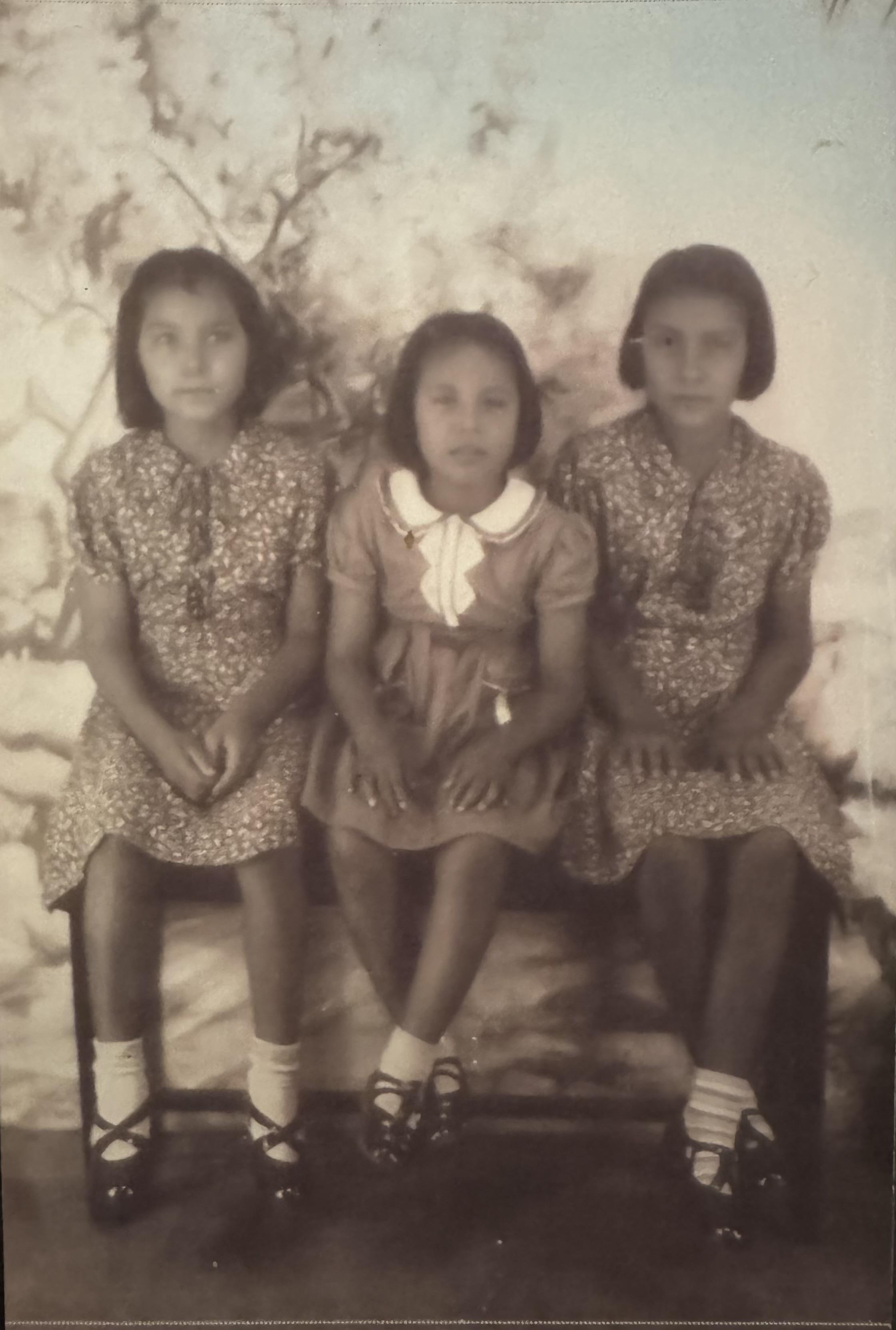Three young girls are seated closely on a bench, wearing floral dresses and smiling softly.
