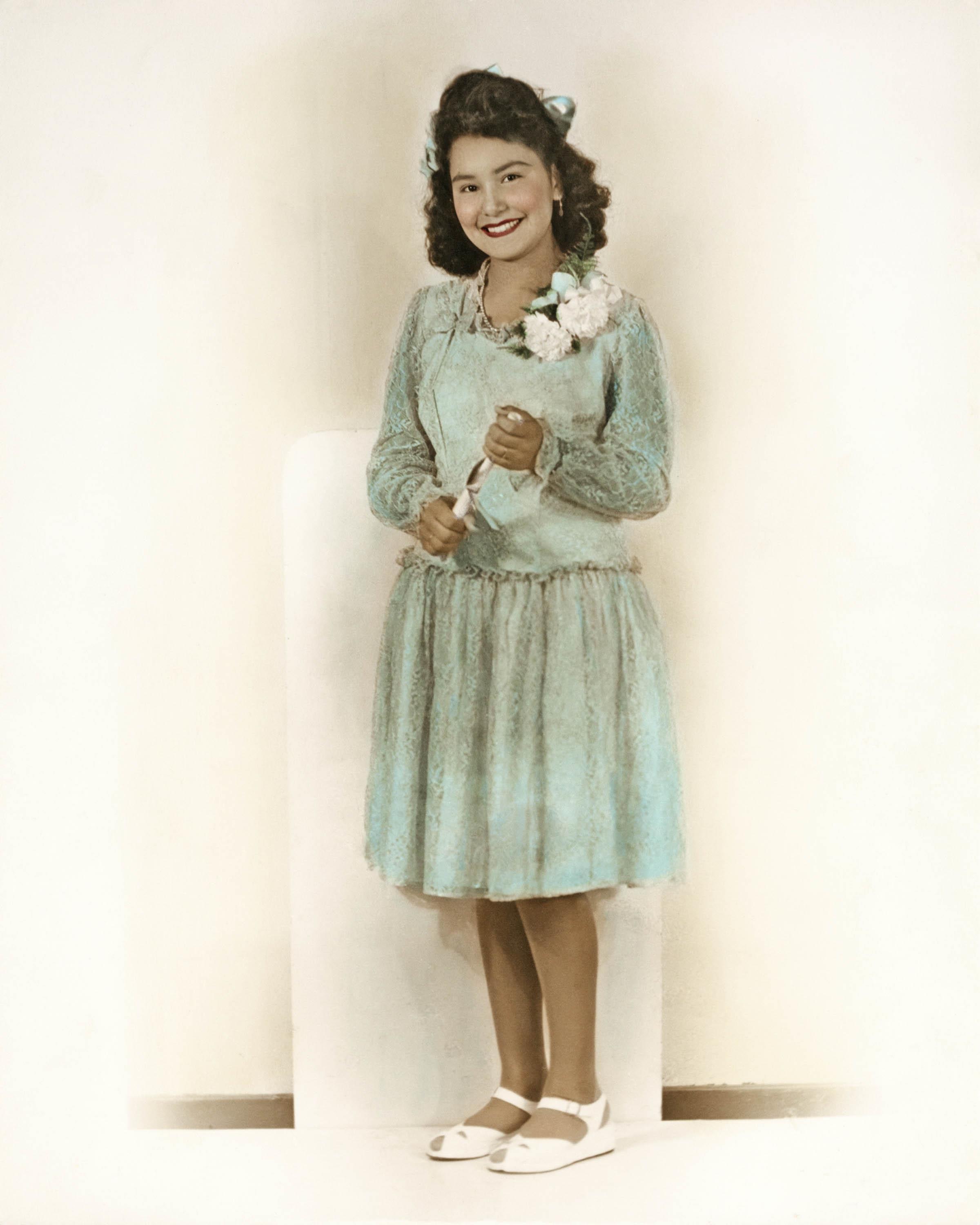 A young girl stands happily in a light blue dress, holding a flower with a big smile.