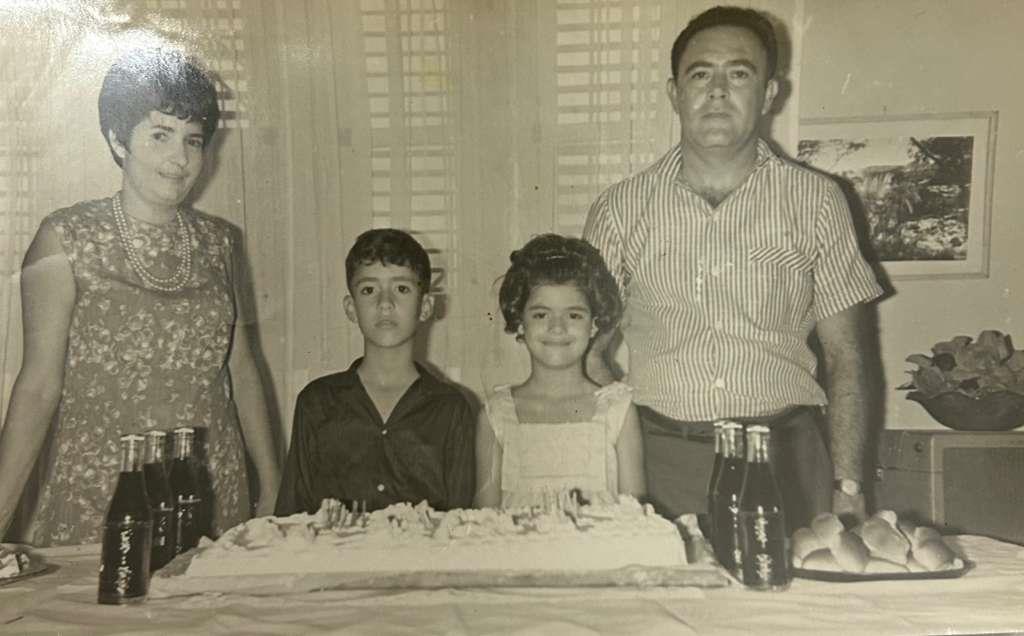 Family gathers around a decorated cake with soft drinks at a home party in the 1960s.