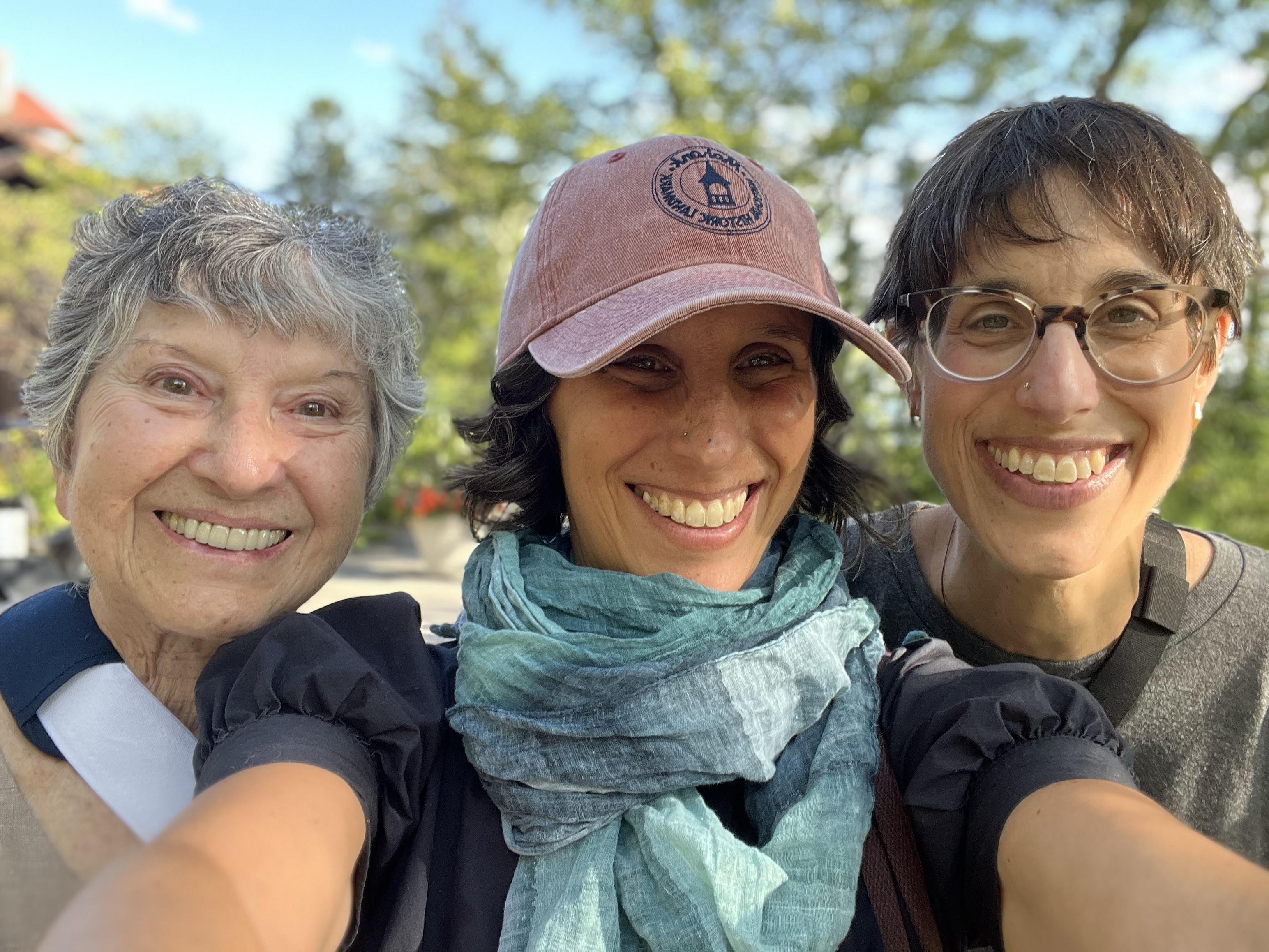 Three women smile happily while taking a selfie in a green park under a blue sky.