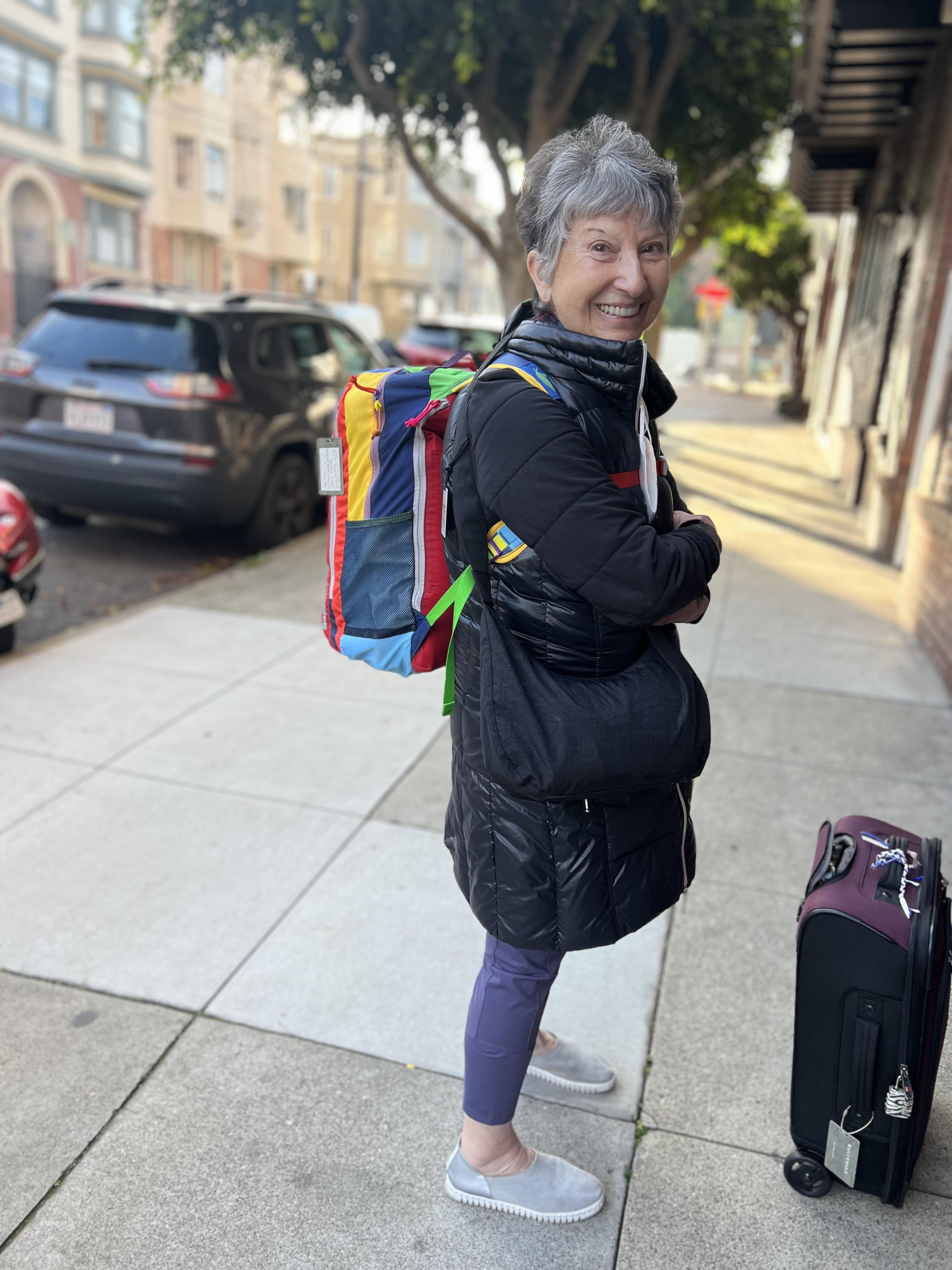 Happy woman stands on a sidewalk with a colorful backpack and suitcase during daylight.