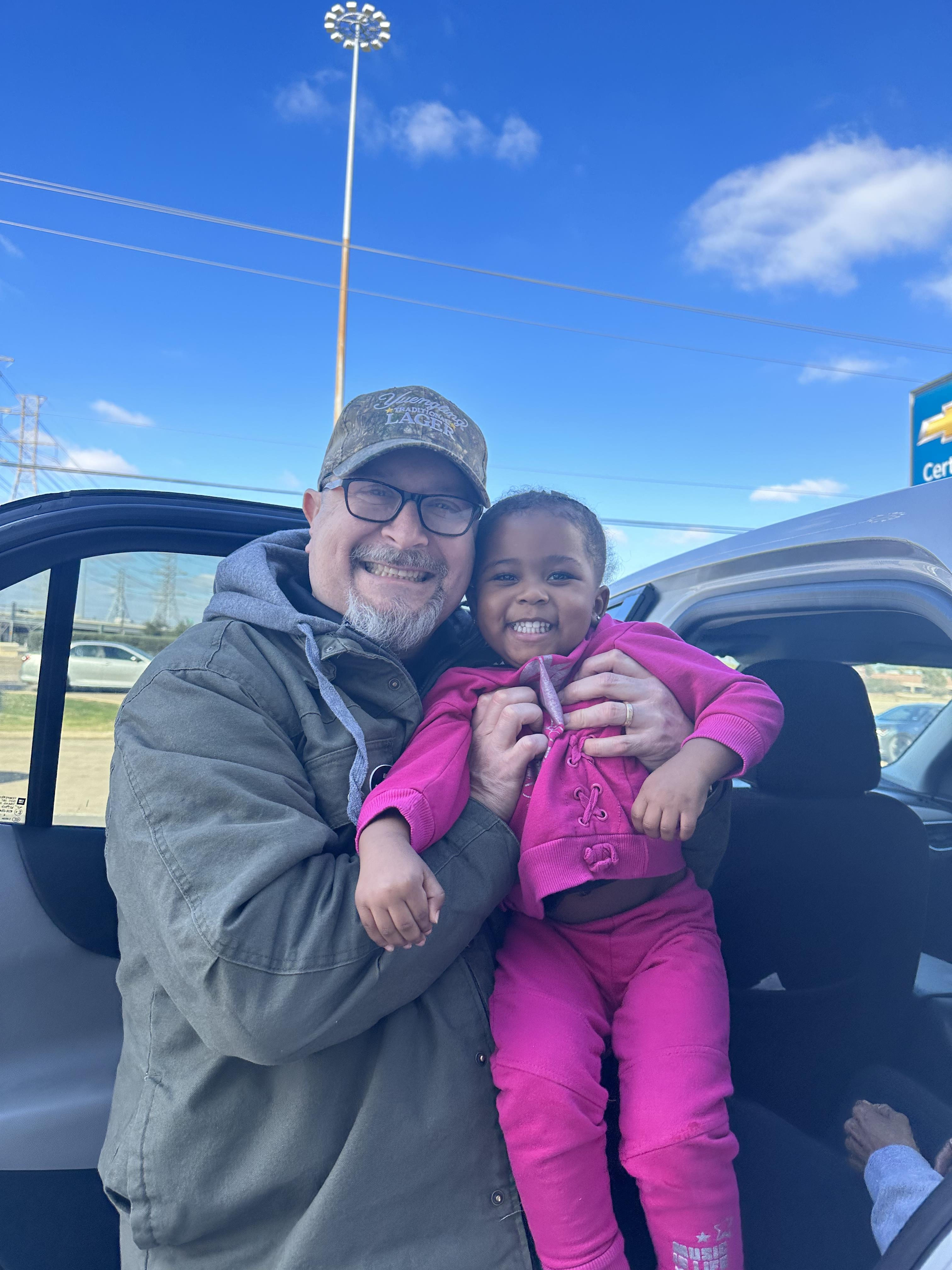 A cheerful grandfather holds his smiling granddaughter in front of a parked car under a blue sky.