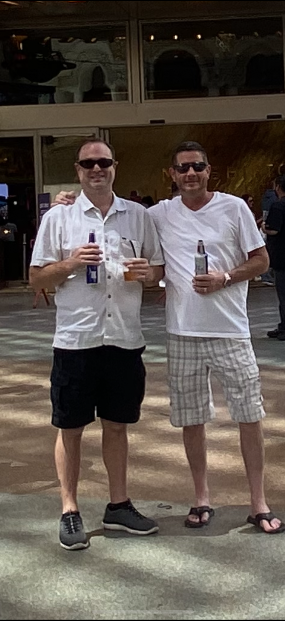 Two friends stand together holding beverages, smiling in a lively outdoor event during the day.