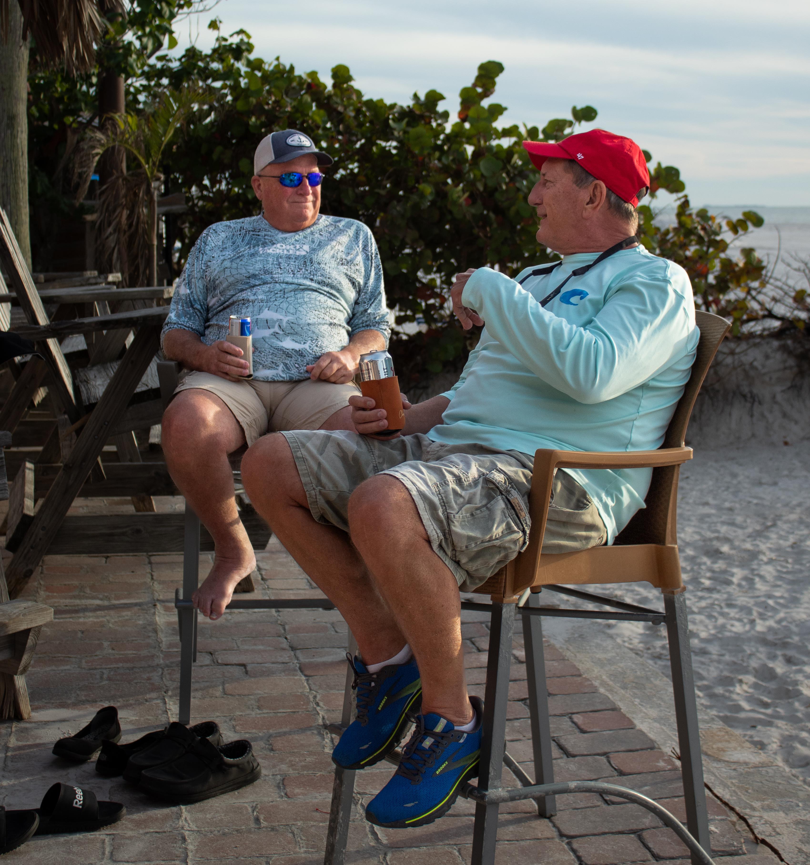 Two men relax on chairs, sipping drinks while chatting by the beach during sunset.