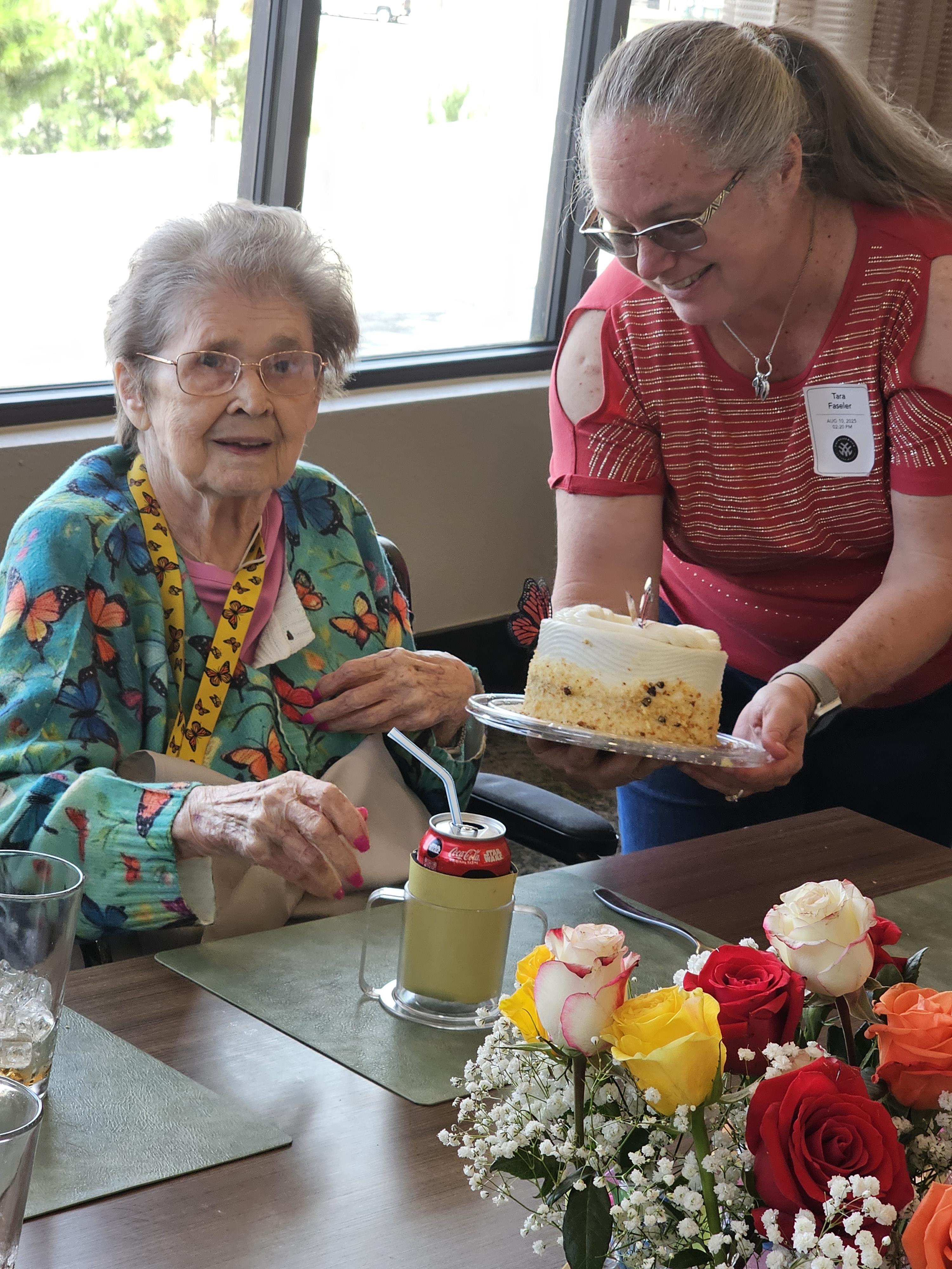 A woman is joyfully surprised by a cake amid a flower-filled birthday celebration.