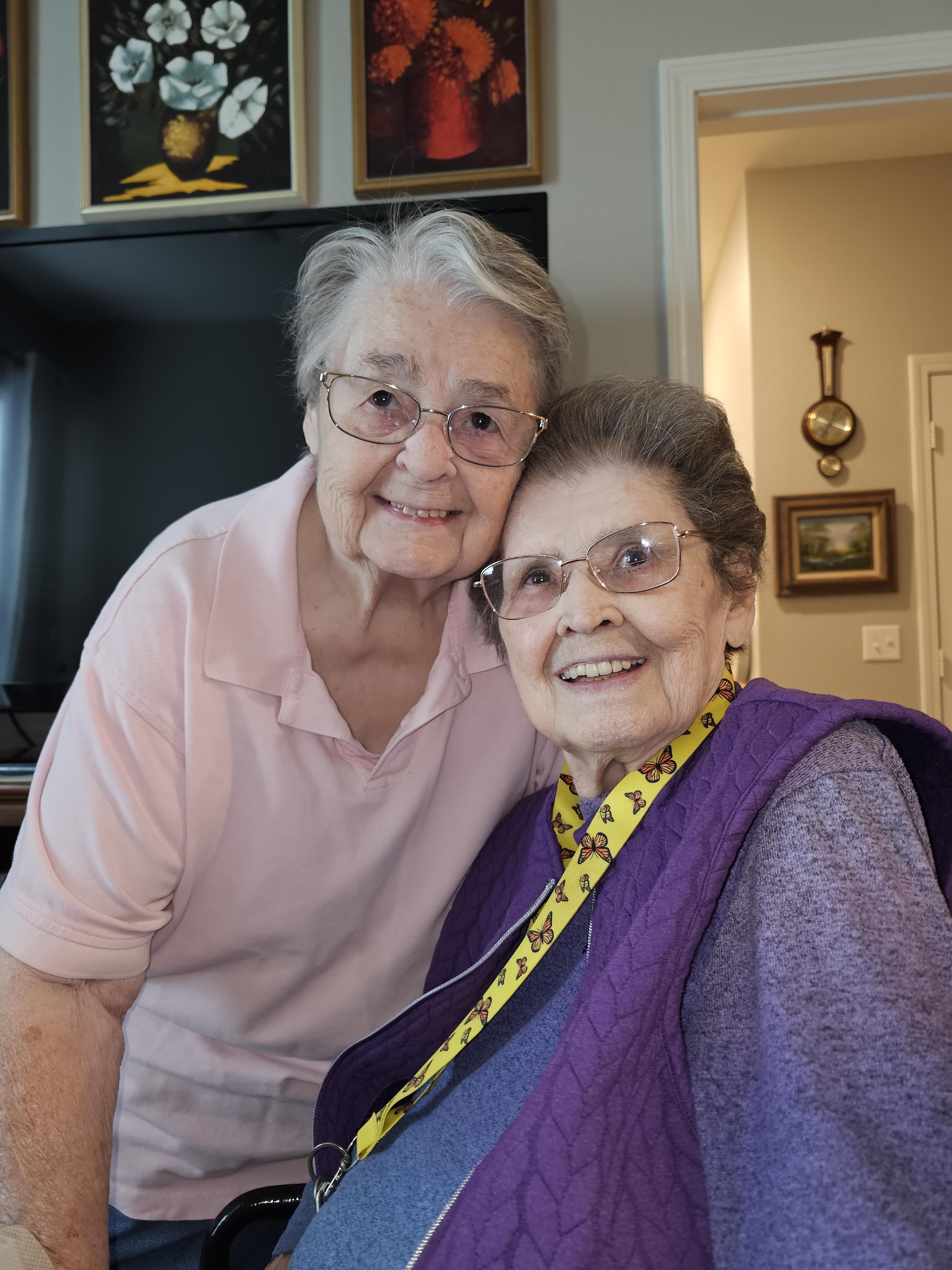Two smiling elderly women pose closely together, sharing a warm moment in a cozy living room.