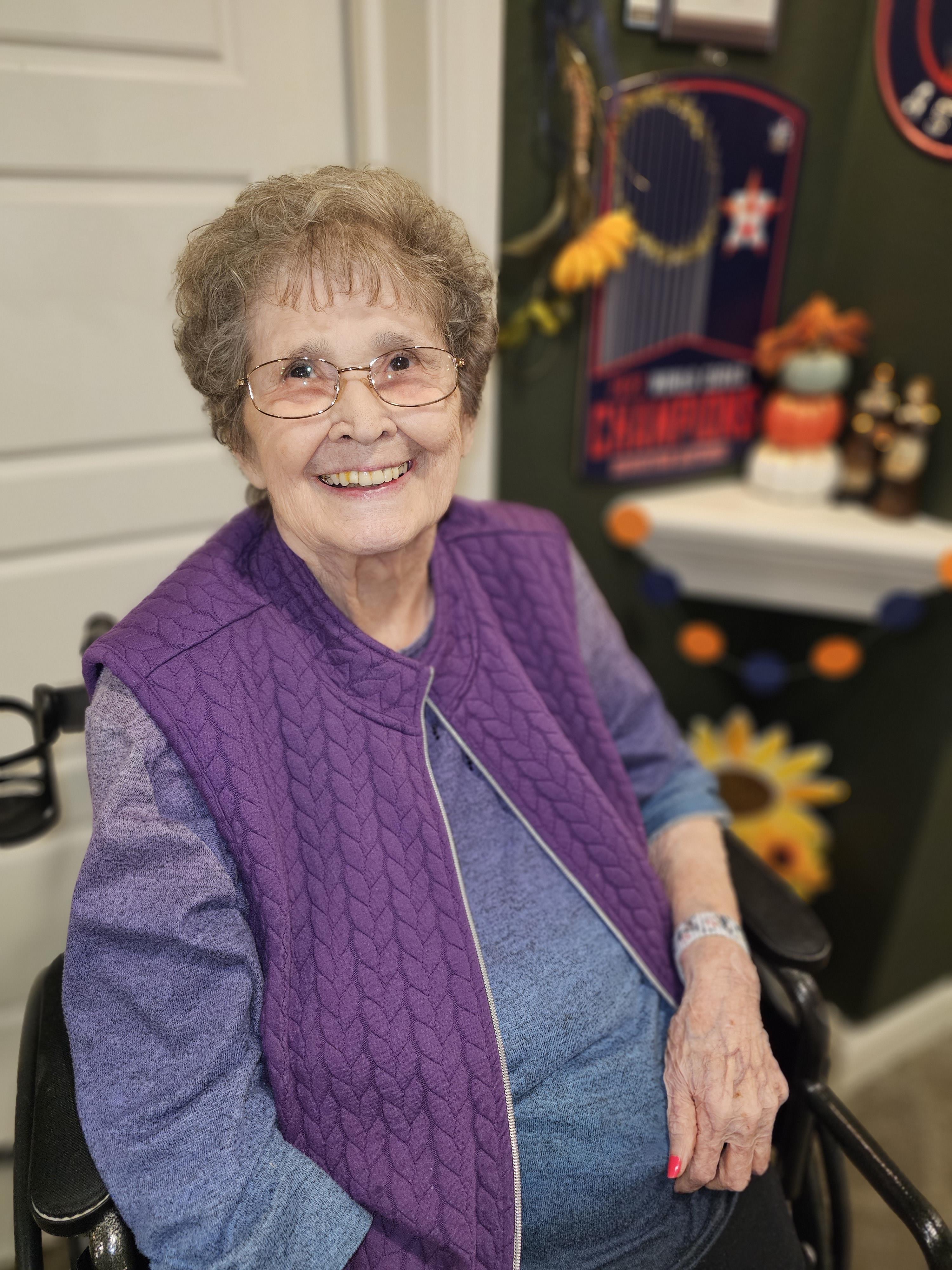 A cheerful elderly woman in a wheelchair, wearing a purple vest, is surrounded by autumn decor.