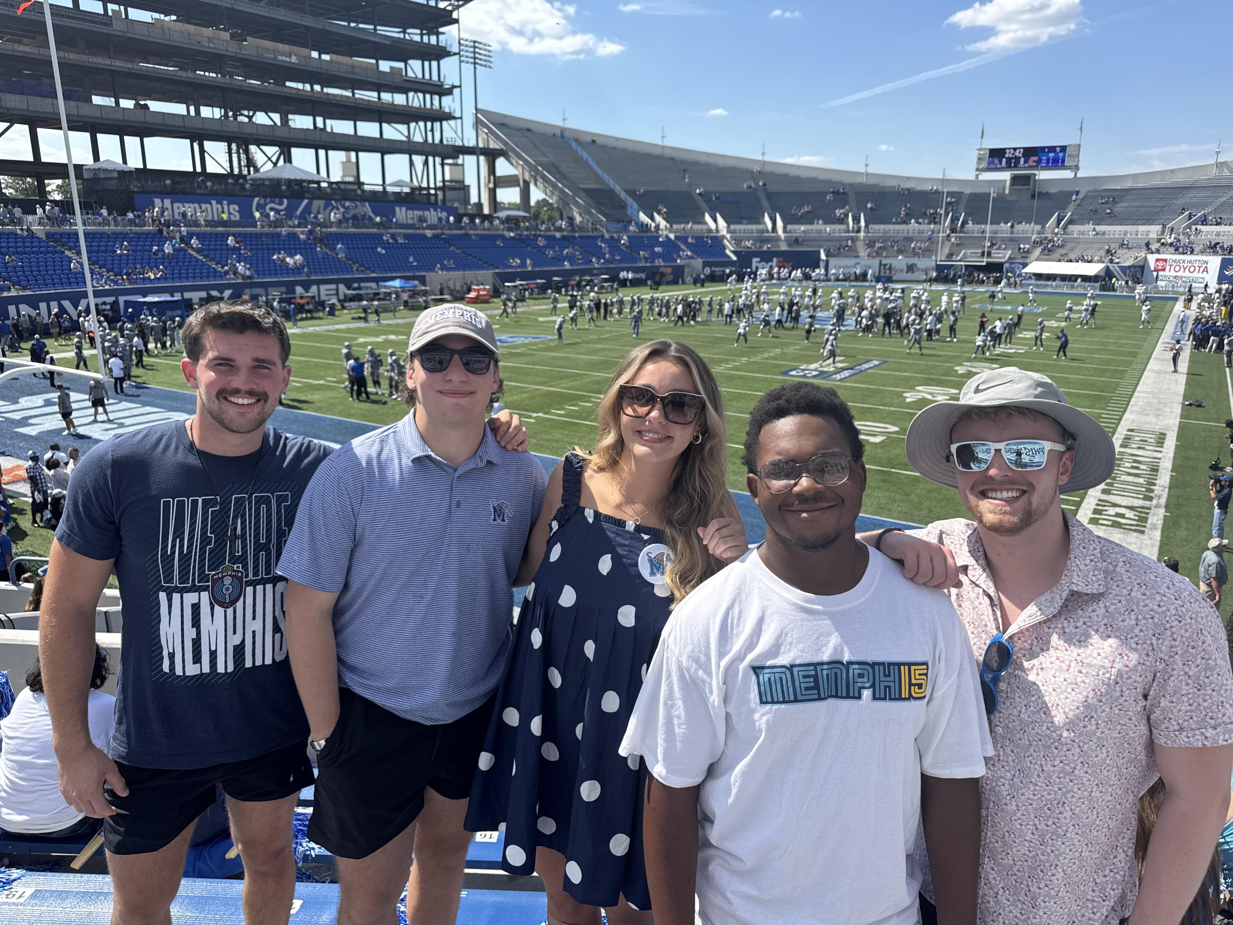 Friends are gathered together smiling at a football stadium before a game on a bright day.