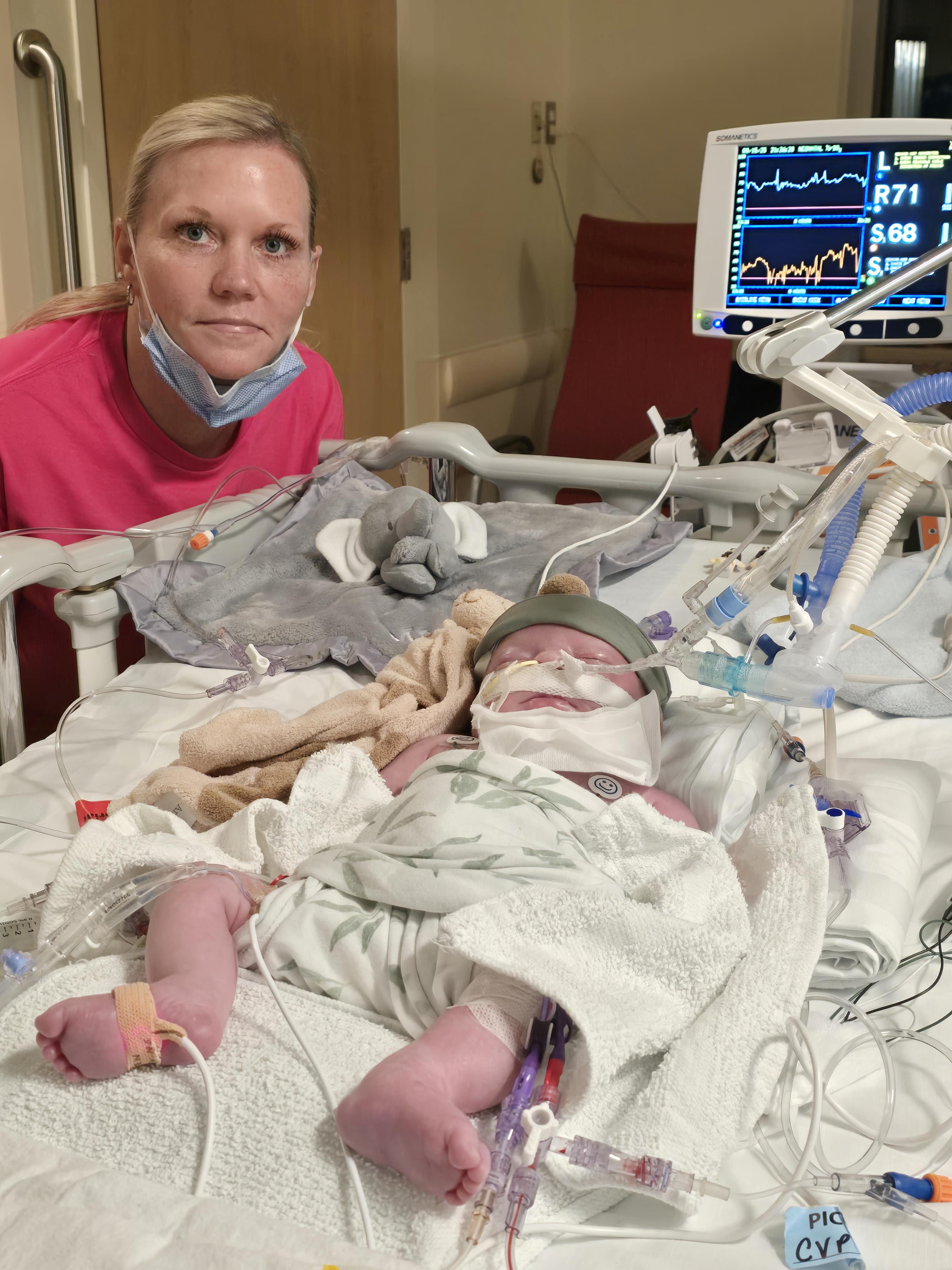 A mother watches over her newborn in a hospital bed surrounded by medical equipment.