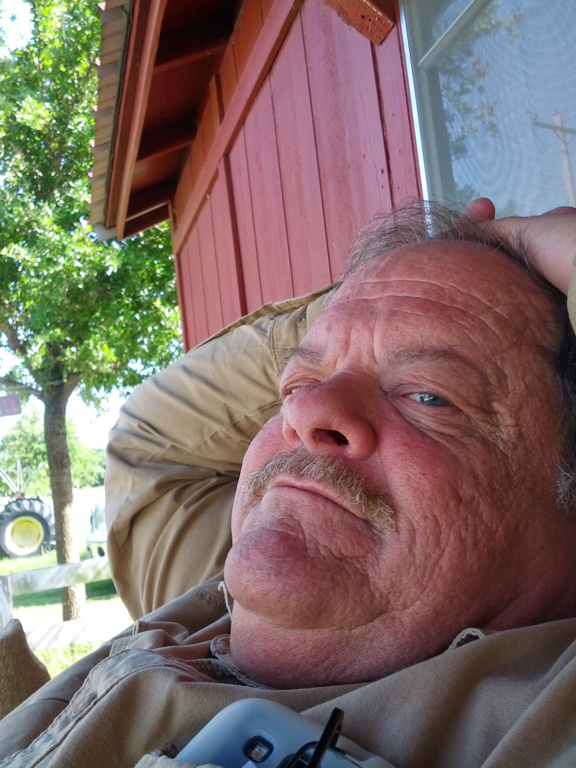 An older man with a relaxed expression enjoys the outdoors, leaning against a wooden wall.