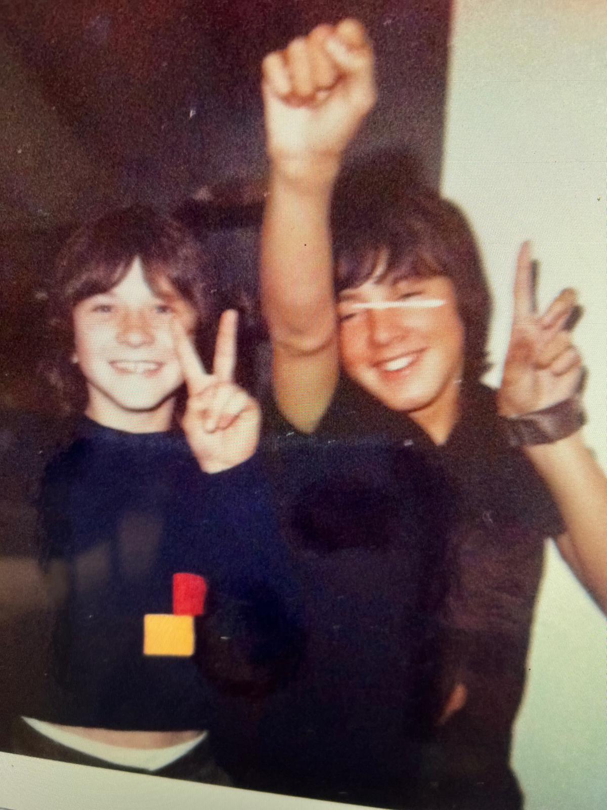 Two kids happily pose with peace signs, enjoying a fun moment together indoors.