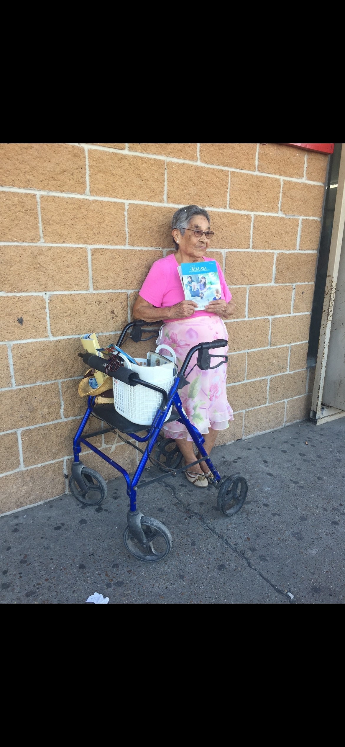 A senior woman in a pink top leans against a wall holding a book while using her walker.