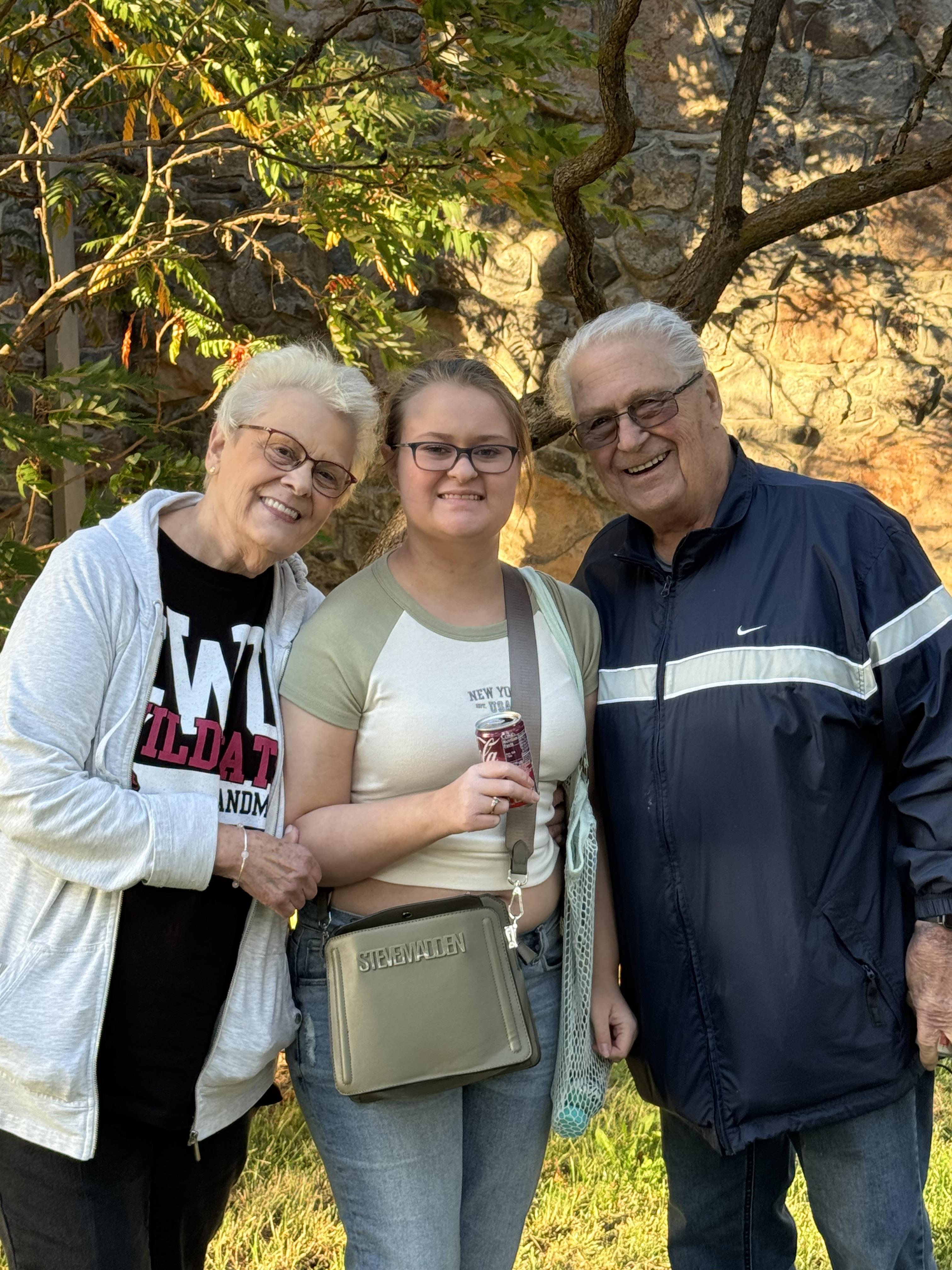 Grandparents stand close to their granddaughter while smiling and posing outdoors during daytime.