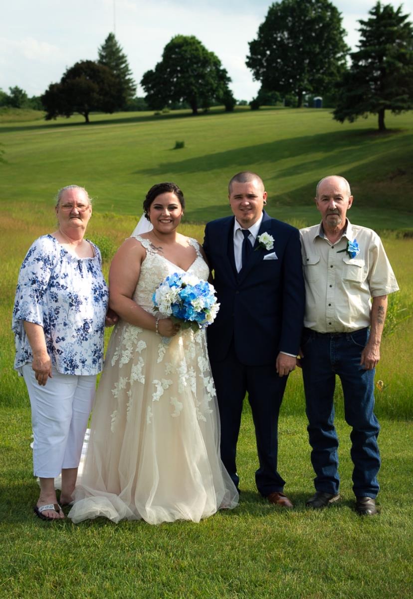 A happy bride and groom pose with their parents on a sunny day in a lush green field.