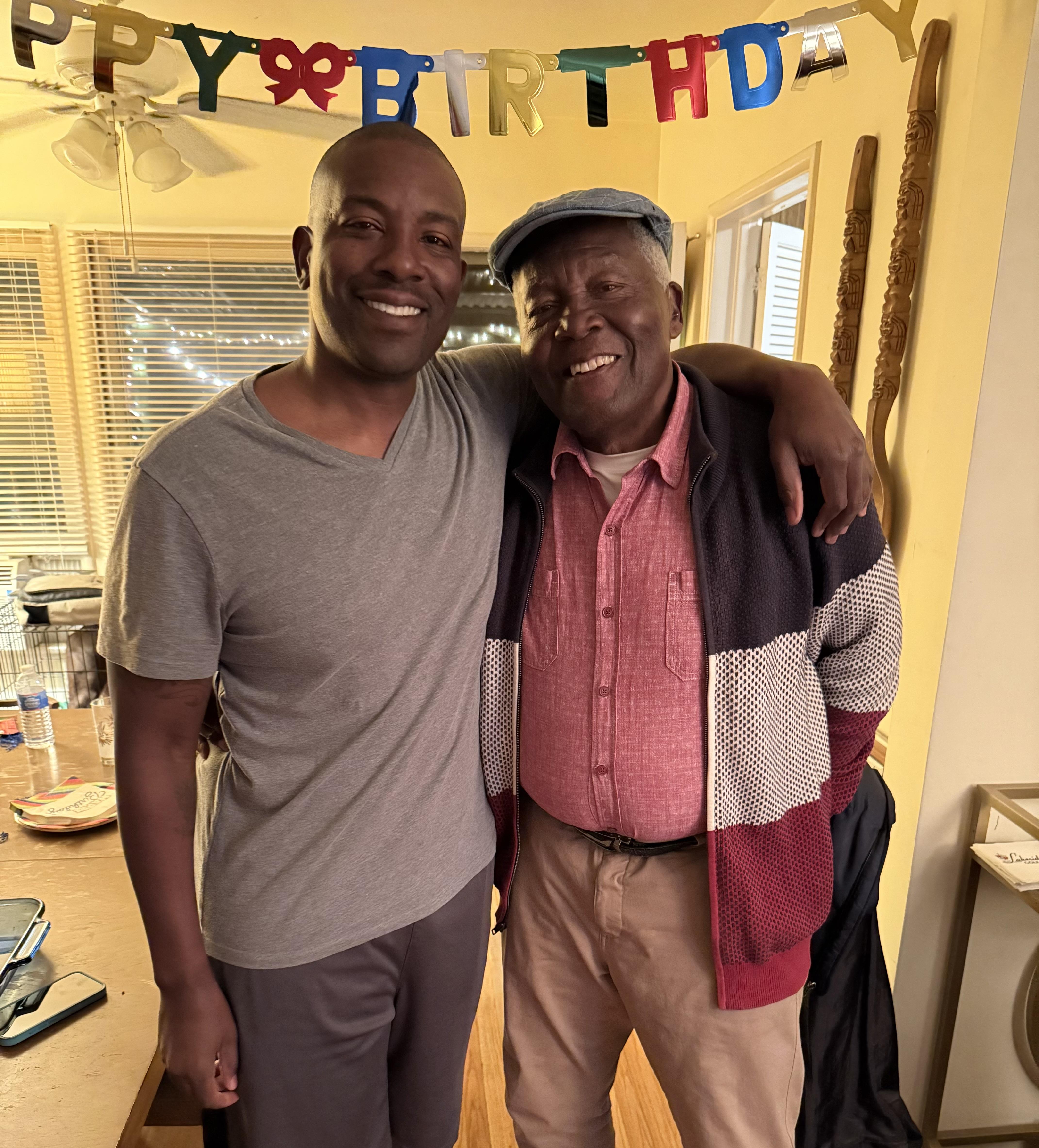 Two men stand close together smiling at a birthday party taking place inside a home.